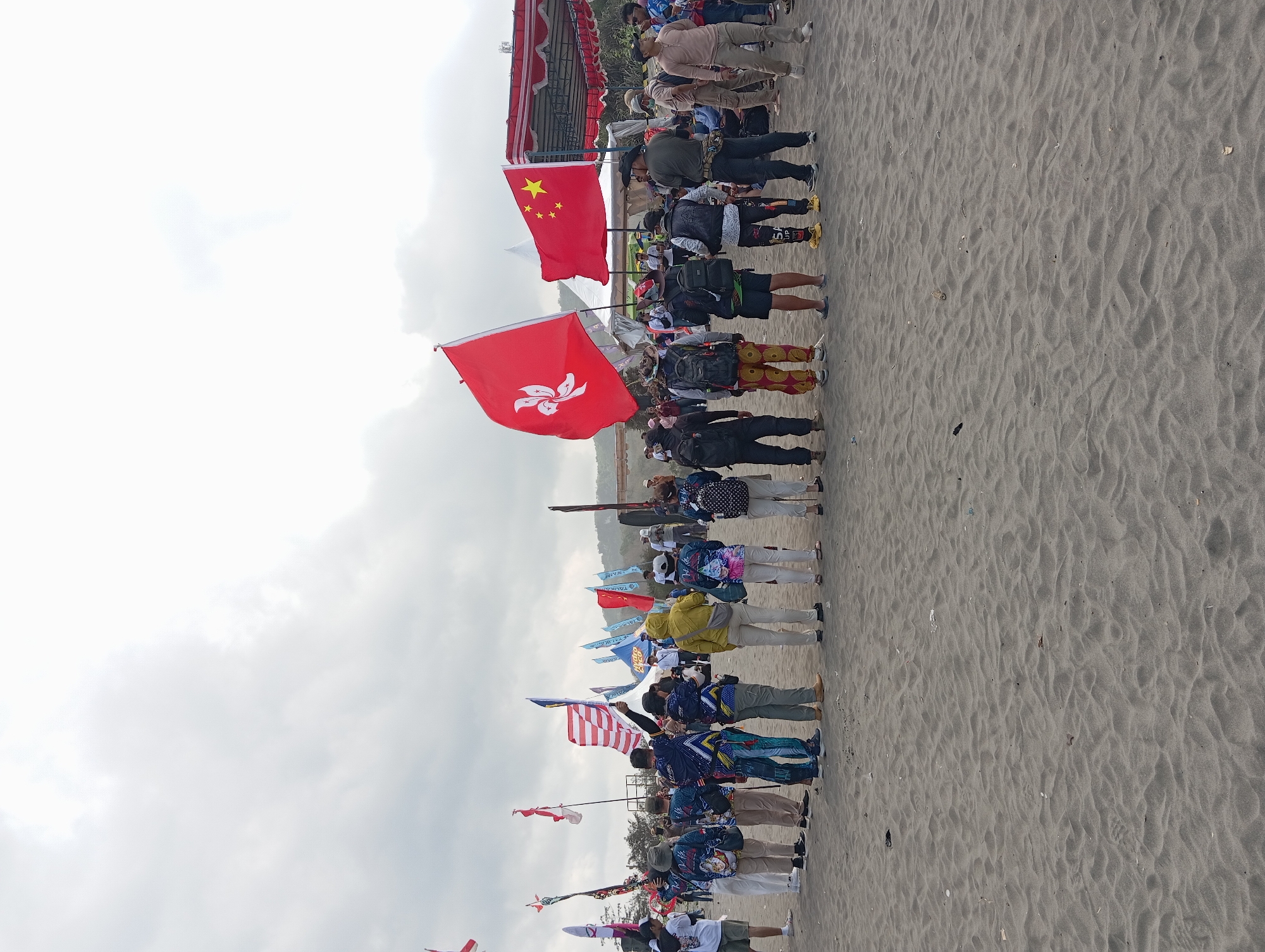 The image depicts an outdoor gathering on a sandy beach, likely in Parangtritis, Indonesia, given the context. A group of people, dressed in casual attire, are standing in a line facing away from the camera, towards a scene with flags and tents. Several individuals are holding flags. Prominently displayed are the flags of Hong Kong and China. The ground is covered in dark sand, and the sky is overcast with gray clouds. In the background, there are tents, a stage-like structure with a red and white canopy, and more flags on poles. The overall atmosphere appears to be that of an organized event or festival.