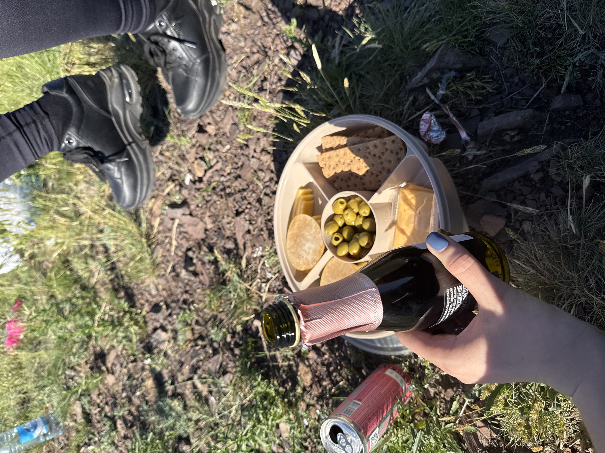An overhead shot depicts a casual outdoor picnic or gathering. Two individuals are present, evidenced by their feet clad in black leggings and sturdy black boots. A hand, adorned with light blue nail polish, holds a dark bottle of sparkling wine, its neck wrapped in pink mesh. Visible Cyrillic text on the label reads "ИГРИСТОЕ ВИНО СЛАДКОЕ" (sparkling wine sweet), indicating a sweet sparkling wine.
A round, segmented food platter takes center stage, offering crackers, green olives, and cheese cubes, suggesting a light snack assortment. A red aluminum can, bearing partial Cyrillic text "КОЛА" and "ENERGI", lies on the ground next to a clear plastic water bottle.
The setting is an outdoor, unpaved area with a mix of green grass, dirt, and small rocks. Bright sunlight casts distinct shadows, indicating daytime. Minor litter, including a blue wrapper, is visible on the ground. The scene portrays a relaxed outdoor social gathering in Ust'-Abakan, Russia.