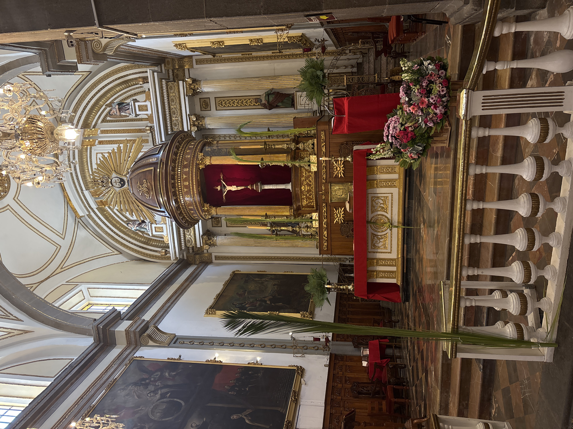 The image shows the interior of a church, presumably located in Puebla, Mexico, judging by the architectural style common in the region. The altar is prominently featured, adorned with gold accents and a crucifix, draped with red cloth. Floral arrangements decorate the front of the altar. Palm branches, possibly for Palm Sunday, are placed near the staircase. The church has grand vaulted ceilings, detailed with gold trim and ornate designs. Large paintings are hung on the walls. A chandelier hangs from the ceiling. The architecture indicates a baroque or neoclassical style, common in historical churches. There is no people in the view.