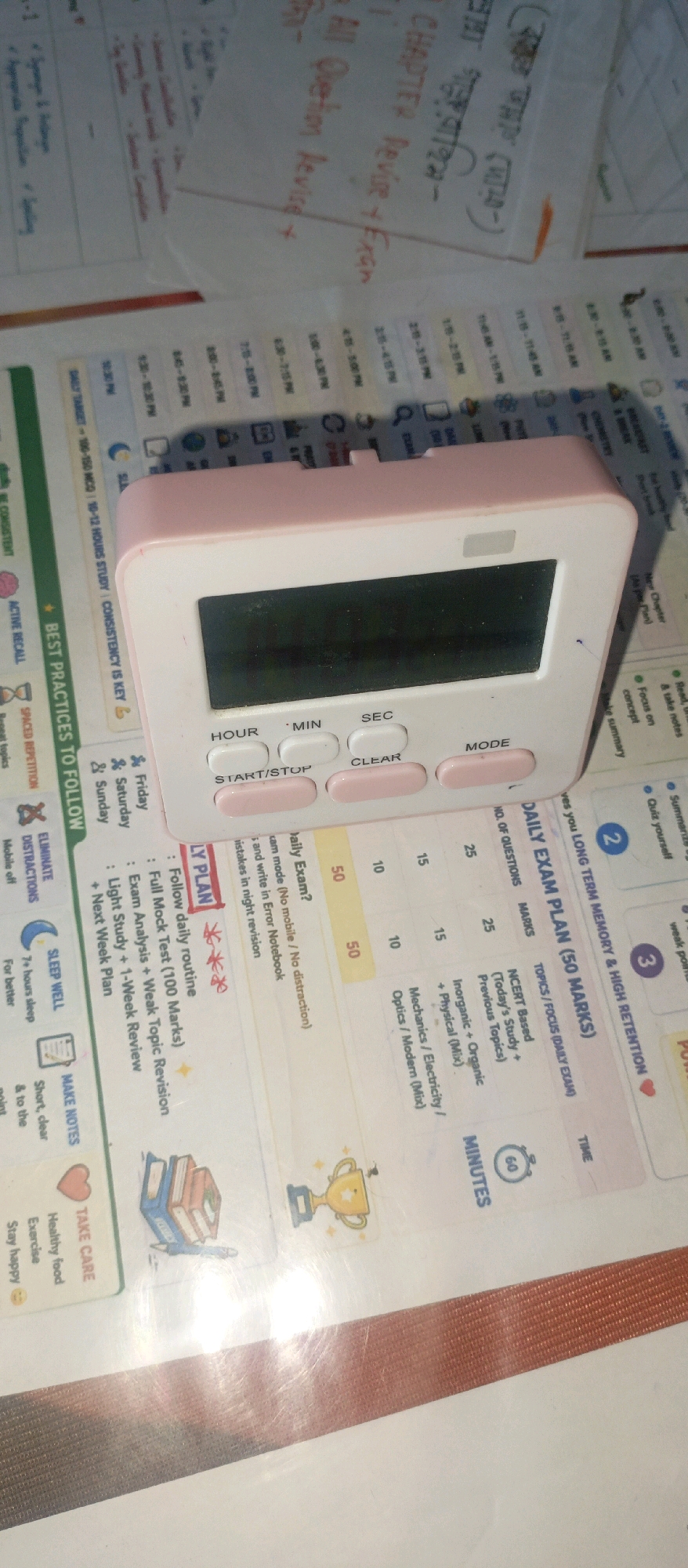 This is a close-up, top-down, angled photograph of a pink digital timer placed on a white and yellow printed planner. The planner has sections labeled "DAILY EXAM PLAN (50 MARKS)" and "BEST PRACTICES TO FOLLOW." A handwritten note in red ink is visible on the planner, reading "DAILY PLAN" followed by bullet points. The timer has a digital screen and several buttons labeled "HOUR," "MIN," "SEC," "START/STOP," "CLEAR," and "MODE." The background shows a blurry assortment of papers and a laminated surface, with some handwritten text on the papers in what appears to be Bengali. The scene is indoors, on a flat surface. There are no people visible in the image. The lighting suggests a well-lit indoor environment. There is no information to suggest the image was taken in Gazipur, Bangladesh.