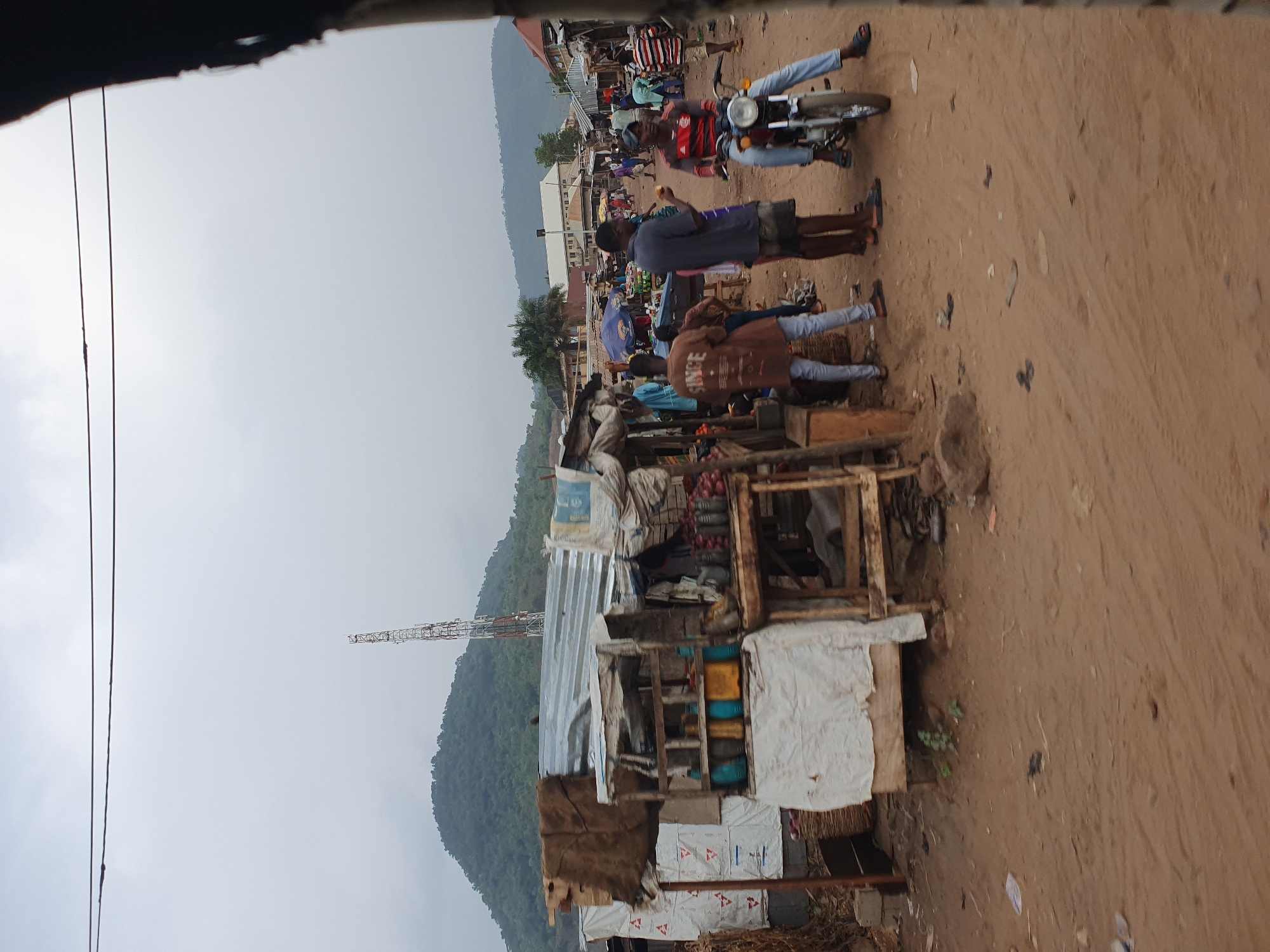This image depicts a bustling outdoor scene, likely a roadside market or commercial area in Lokoja, Nigeria. The setting features a wide, unpaved dirt road that serves as the main thoroughfare. In the background, lush green hills or mountains rise under an overcast, grey sky, indicating a cloudy day with soft, diffused lighting. A tall communications tower is visible on the left side of the distant mountainside.
Several people are present, engaged in various daily activities. In the midground, a man sits astride a motorcycle, wearing a red and black striped shirt and blue jeans. Nearby, another man in a dark blue shirt and shorts stands, holding a piece of fruit, possibly an orange. Further into the scene, other individuals are walking or gathering, some in proximity to a makeshift stall. One person wears a brown jacket with the word "SINCE" visible on the back.
On the left, a rudimentary stall or shop is constructed from wooden planks, corrugated metal sheets, and tarpaulin or sacking materials, suggesting an informal business setup. Various goods are visible within and around the stall, including sacks of items, bottles, and what appears to be produce, such as onions in a basket. Overhead, utility lines or power cables stretch across the upper left corner of the frame. The overall atmosphere conveys a typical day of commerce and community interaction, with people going about their routines in a lively, semi-urban environment.