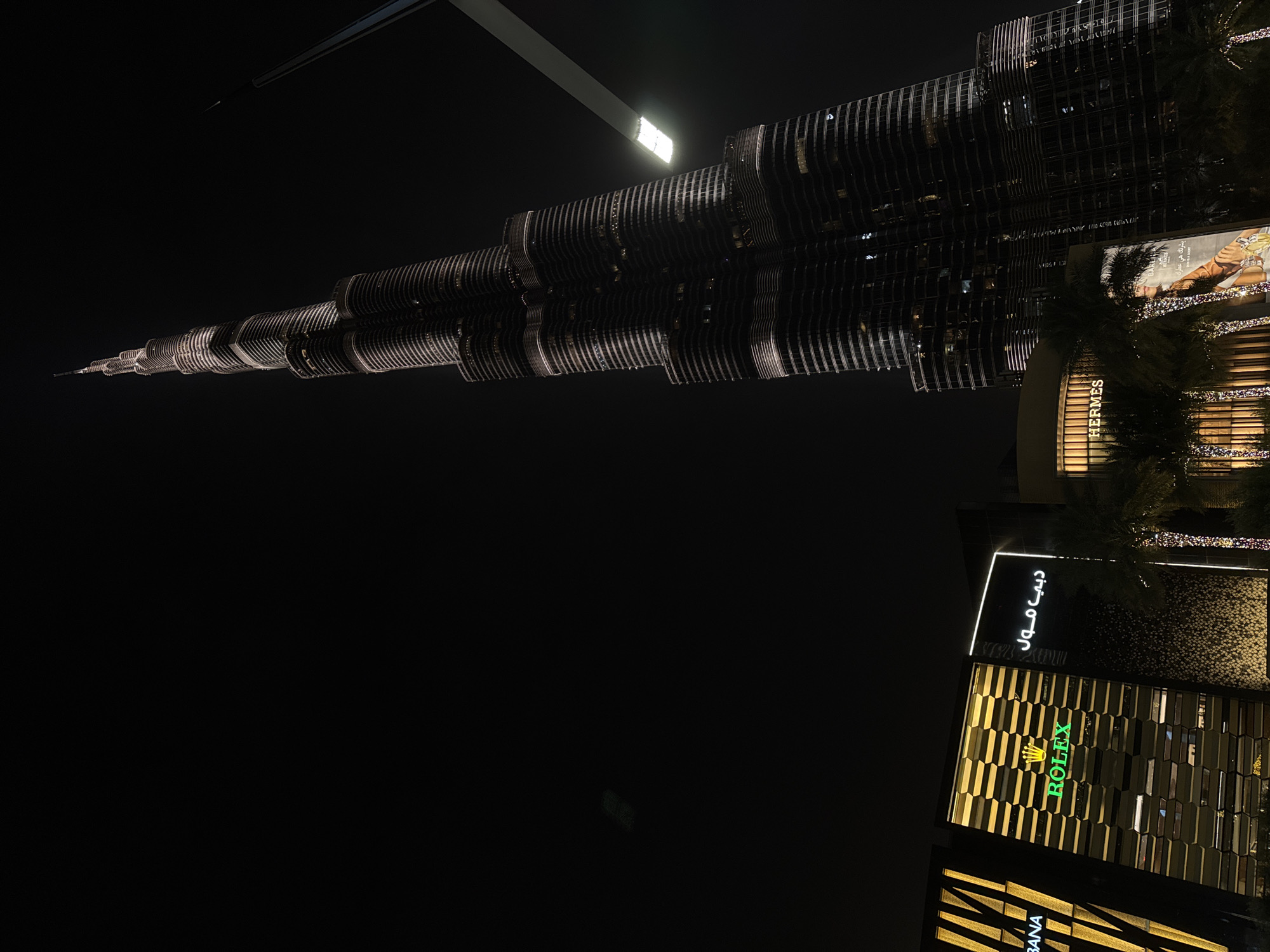 The image shows the Burj Khalifa in Dubai, United Arab Emirates, at night. The skyscraper is brightly lit and dominates the scene. In the foreground, illuminated store signs for Rolex, Dolce & Gabbana, Hermes, and Dubai Mall are visible. Palm trees are scattered around the base of the buildings. The environment is dark, with the sky being black.