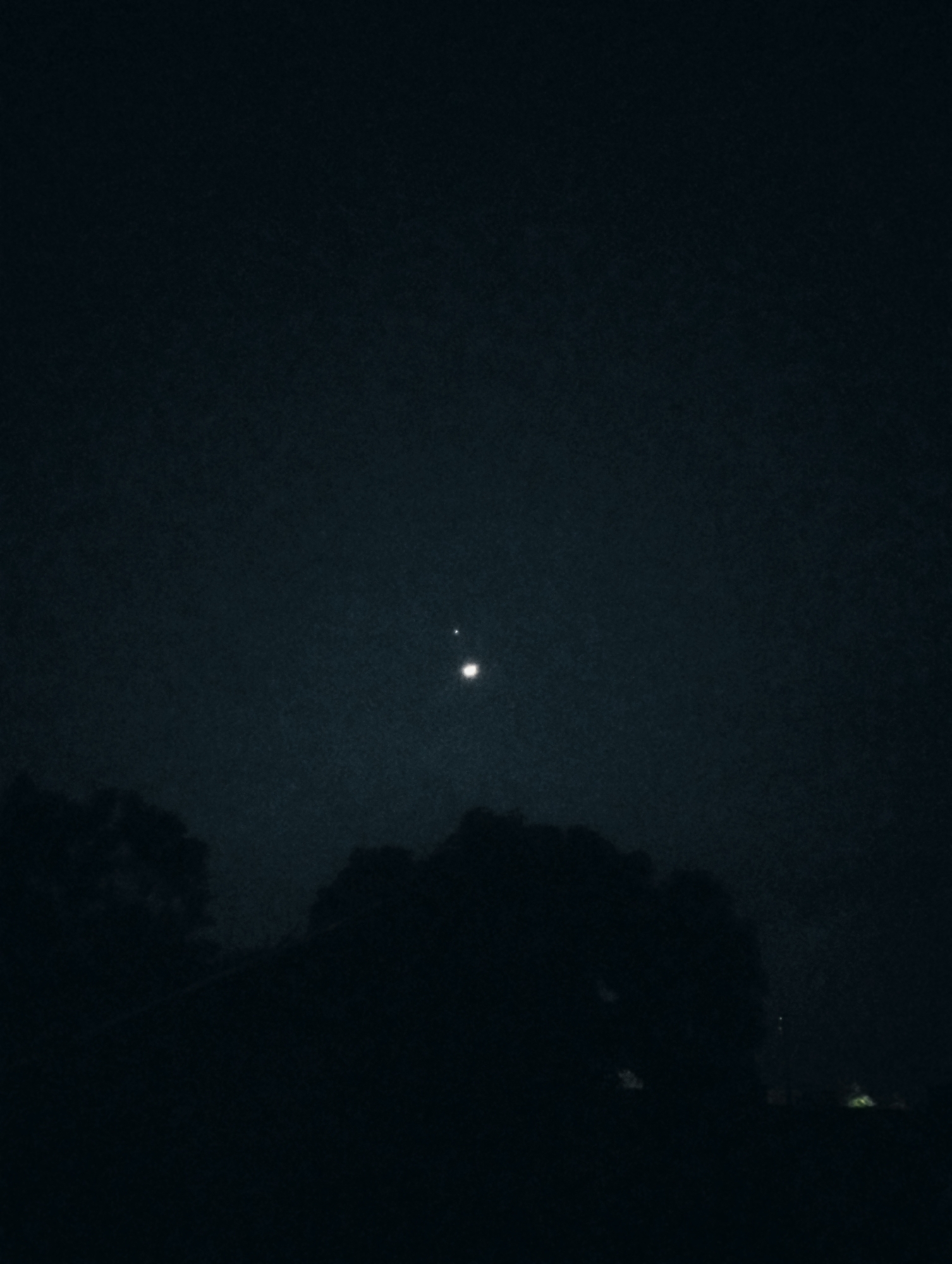 The image shows a dark, nighttime scene with a slightly hazy sky. In the upper half of the image, two bright celestial bodies are visible. The lower half of the image is dominated by the silhouette of trees and what appears to be a streetlamp or distant lights. The scene is devoid of any people. Given the low-light conditions and the presence of what appears to be a streetlamp, the location could be an urban or semi-urban area. Without further information or distinct landmarks, it is not possible to definitively pinpoint the location within Jos, Nigeria. The overall impression is one of quietness and the vastness of the night sky.