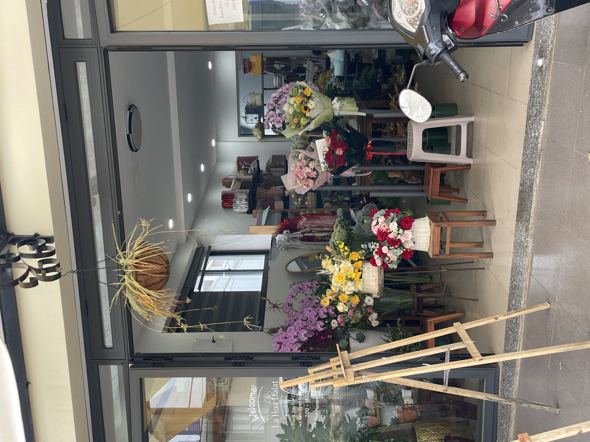 This is a full shot of the exterior of a flower shop named "La Hoa Da Lat" in Lam Vien - Da Lat, Vietnam. The shop entrance is open, revealing a colorful display of flower arrangements and plants. In front of the shop, there's a wooden easel and a few stools. A red and black scooter is parked to the right of the entrance. A hanging basket with a plant hangs above the doorway. The shop appears to be a standalone establishment on a street, with a clean, tiled sidewalk in front.