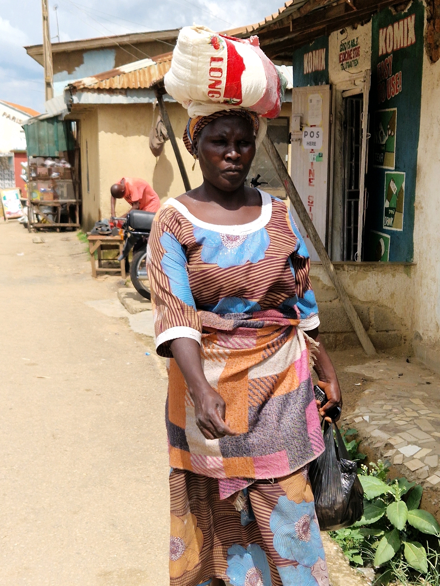 In Kabong, Nigeria, a woman walks down a dirt road carrying a large white sack of goods on her head, with "LONG" written in red letters on the sack. She wears a vibrant, patterned wrap skirt and a striped blouse with blue floral designs. In her right hand, she holds a black plastic bag, and in her left, a mobile phone. Behind her, a person in an orange shirt is hunched over a motorcycle. The scene is set against a backdrop of modest buildings with corrugated metal roofs and colorful signs.