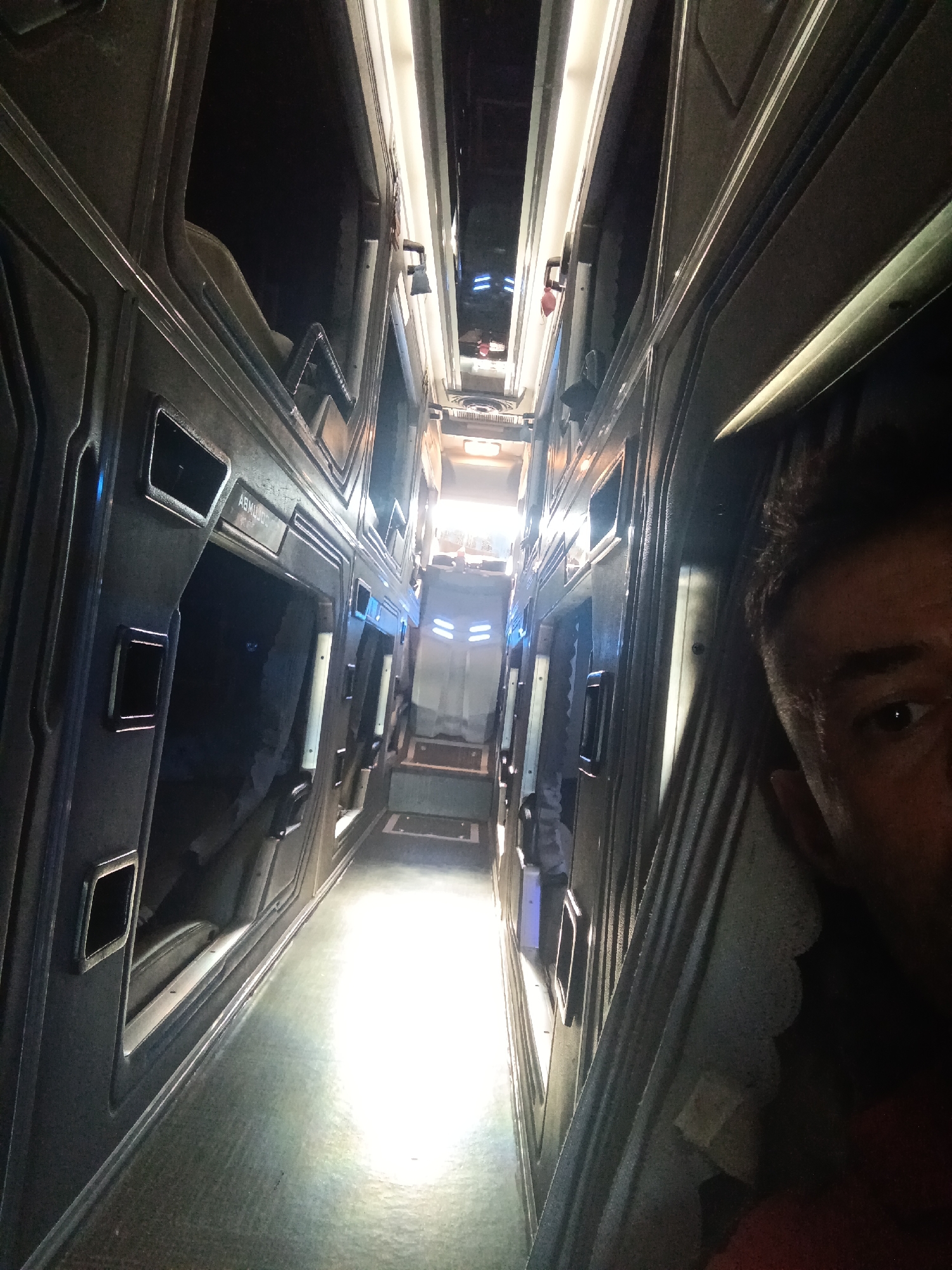 The image depicts the interior of a sleeping pod or capsule hotel hallway, likely in Phu Thuy Ward, Vietnam. The hallway is narrow and lined with rows of sleeping pods on both sides, with a bright, well-lit corridor running down the center. The pods appear to be constructed from dark, metallic-looking material and have individual openings. Some of the pods are open, revealing a glimpse of the interior space. The lighting is a mix of overhead fluorescent lights and the ambient light from the end of the hallway, which appears to be a doorway or exit. In the foreground on the right, the face of a person is partially visible, looking towards the camera. They have dark hair and are looking over their right shoulder. The overall impression is of a functional, modern, and somewhat confined living space.