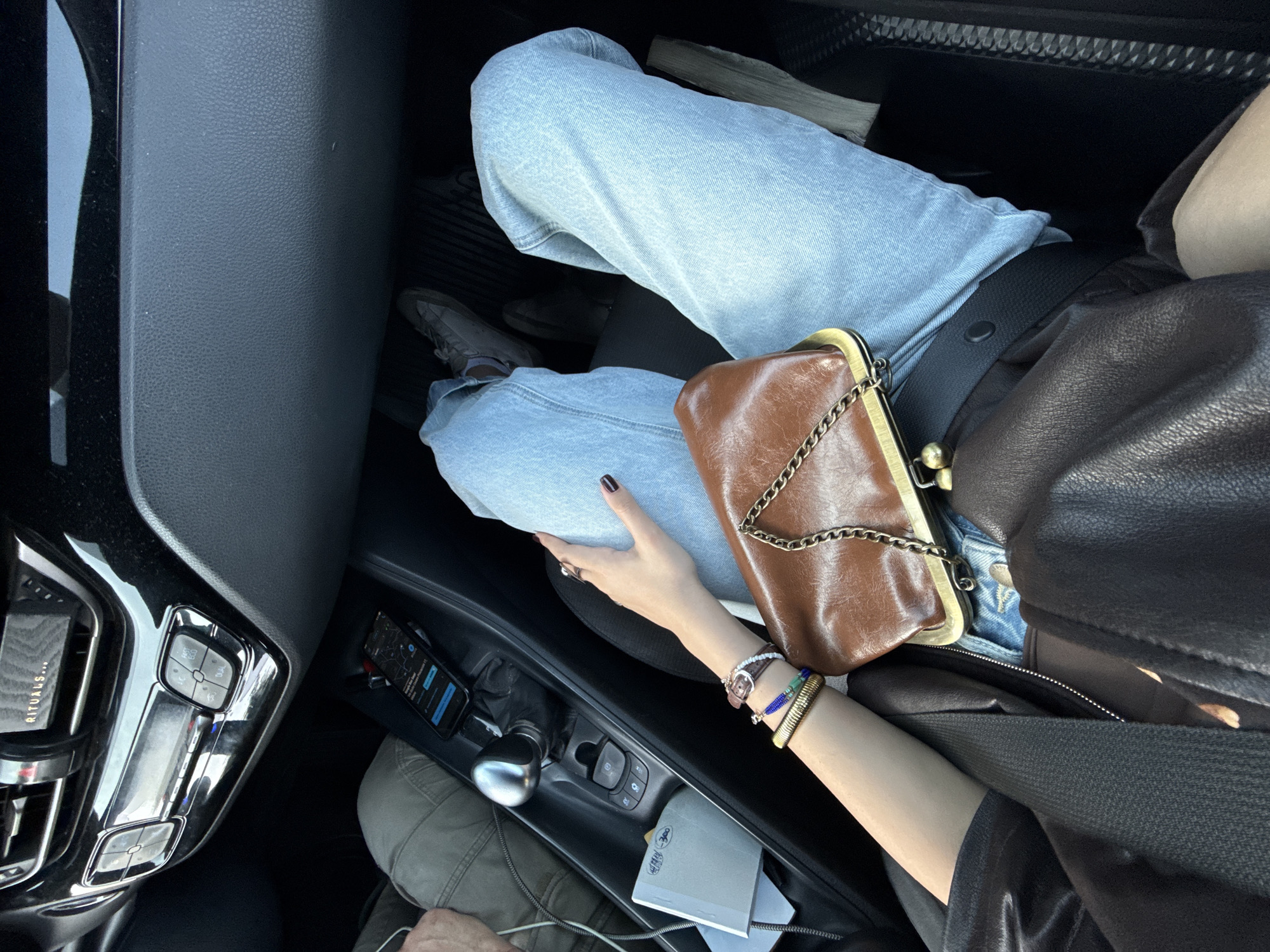 A high-angle shot taken from inside a car. The car's gear shift, adjacent buttons, and a phone with a map on its screen are visible. A person wearing light blue jeans, a black top, and a brown leather jacket is seated in the passenger seat. They are wearing several bracelets on their left wrist. A brown leather purse with a gold chain is resting on their lap. The person is wearing a seatbelt. The location is unknown.