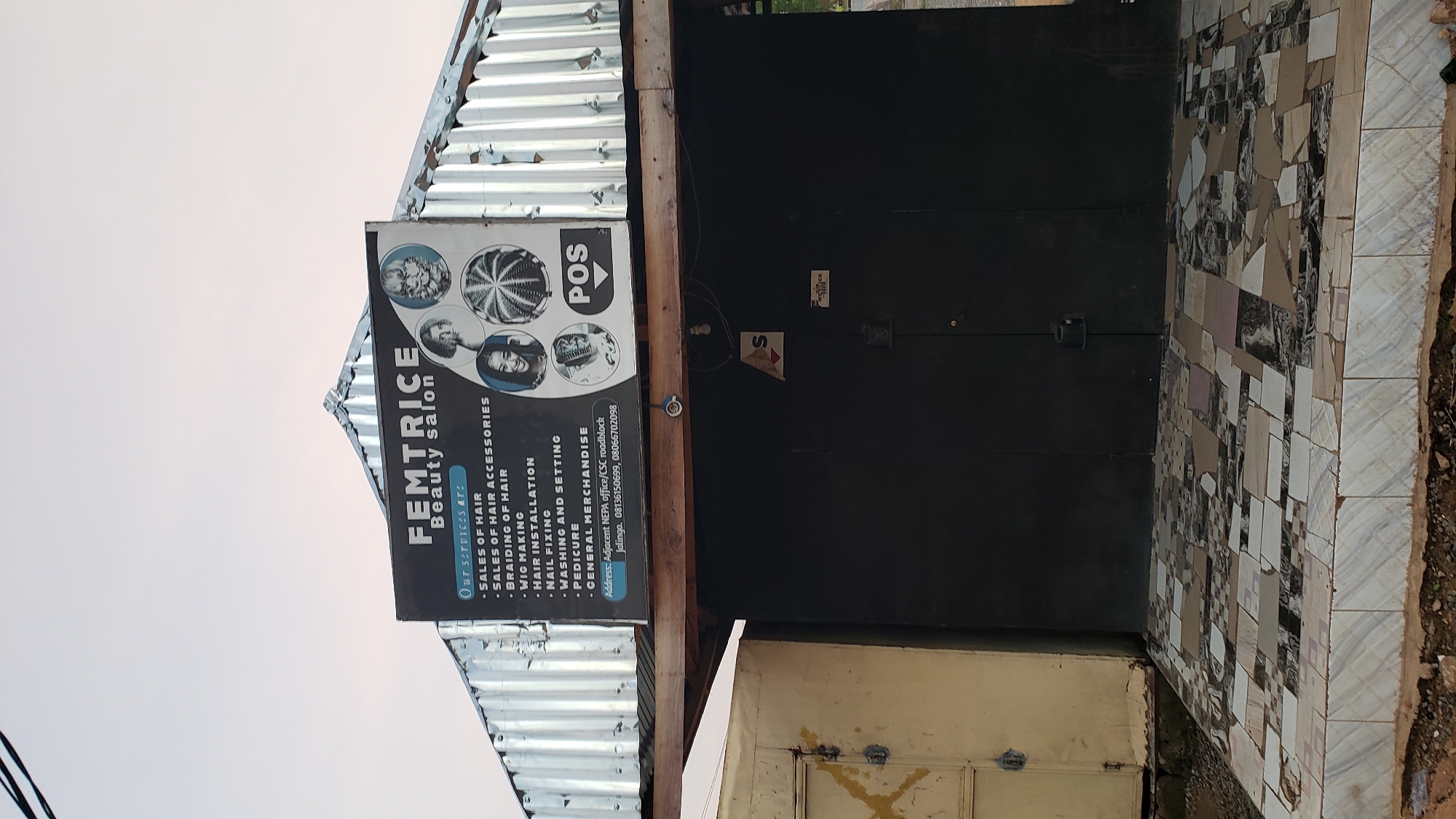 The image shows the exterior of a beauty salon named "FEMTRICE Beauty salon" in Jalingo, Nigeria. The salon is located on the right side of the image, behind a black metal door. Above the door is a sign listing the services offered, including sales of hair, hair accessories, braiding, wig making, hair installation, nail fixing, pedicure, and general merchandise. The sign also provides contact information, with phone numbers 08136150699 and 066702098, and the address "Adjacent NEPA office/CSC roadblock, Jalingo." The salon building has a corrugated metal roof, and the entrance area is tiled with a mosaic pattern. To the left of the salon is a light-colored metal structure, possibly another shop or storage area, with a yellow-painted door. The sky in the background is overcast.