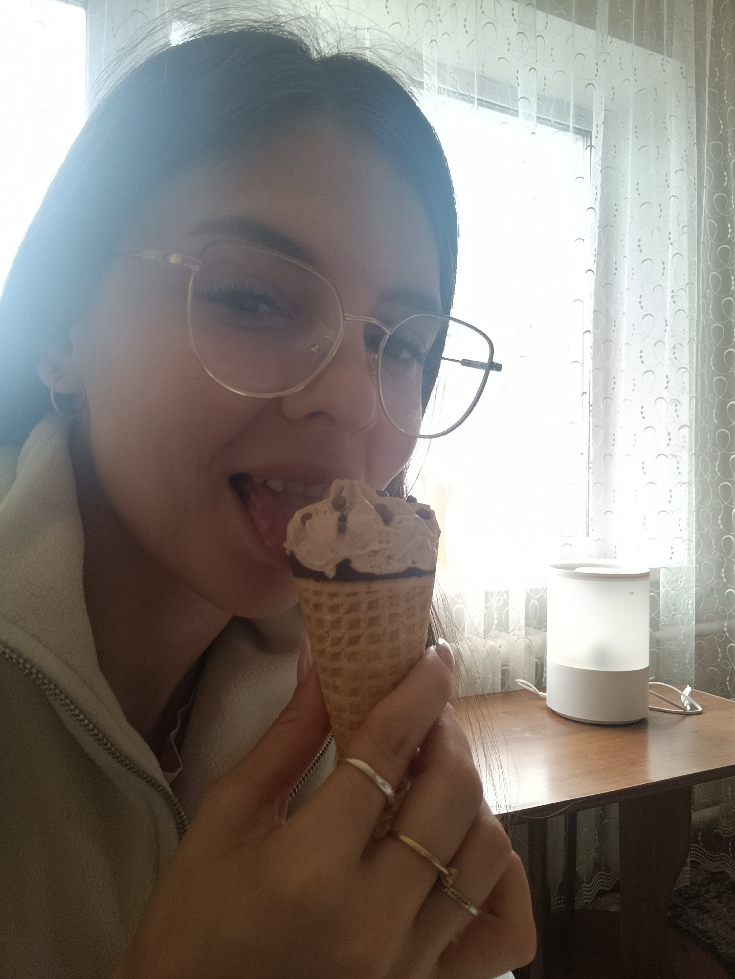 A young woman is holding and licking an ice cream cone indoors. She is wearing glasses and a light-colored zip-up jacket. She has rings on her fingers. A table and a white object (possibly a humidifier or lamp) are visible in the background, along with curtains. The location is unknown.