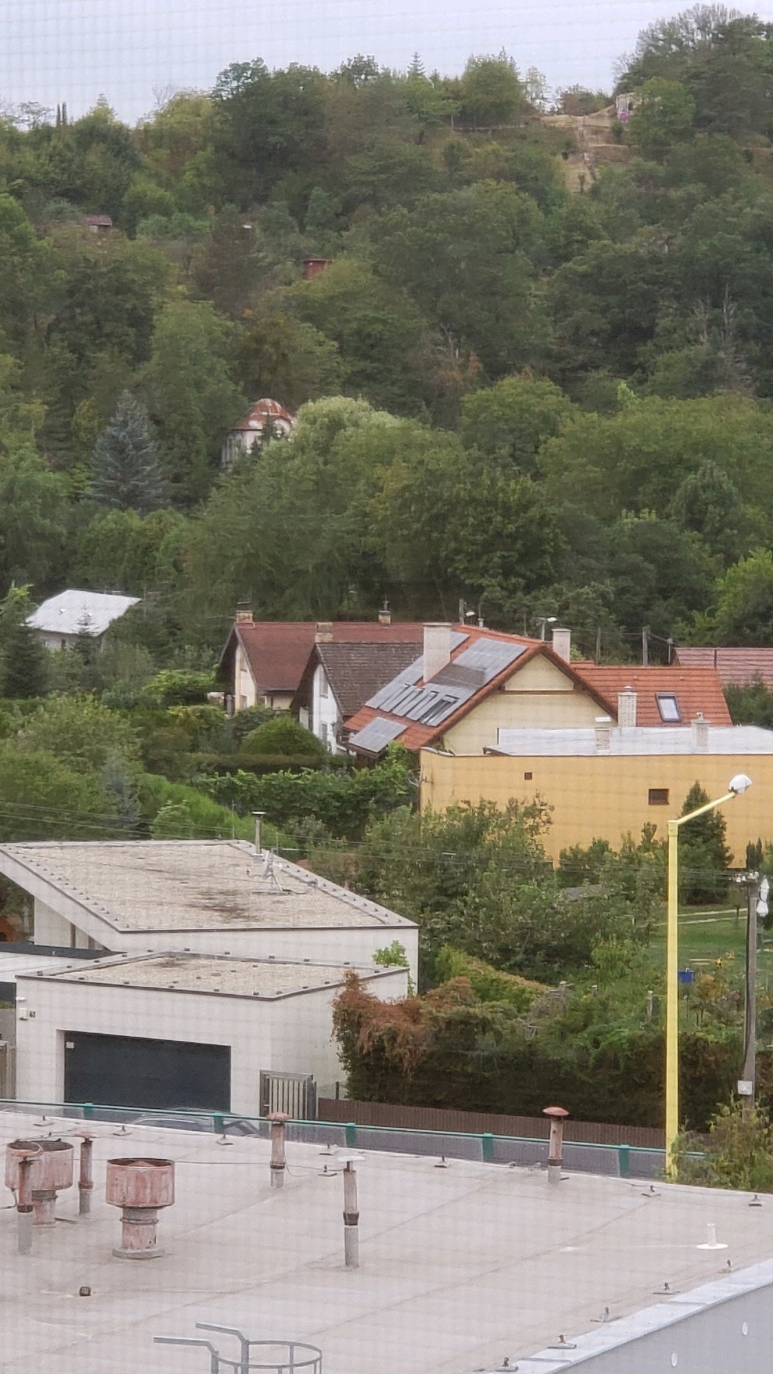 This is a medium-angle shot of a suburban hillside area in Prešov, Slovakia, on an overcast day. In the foreground, the rooftop of a modern building is visible, featuring several ventilation pipes and a light-colored, flat surface. Beyond this, a two-story white building with a dark garage door and a metal fence is partially visible.
Further up the hillside, several houses with red tiled roofs are nestled amongst dense green trees. One of these houses has solar panels installed on its roof. Another house, with a white facade, is visible to the left, also surrounded by greenery. Higher up on the hill, there are more trees, and the faint outline of what appears to be a small building with a domed roof can be seen amongst them. The overall impression is of a residential area integrated with a heavily wooded natural landscape.