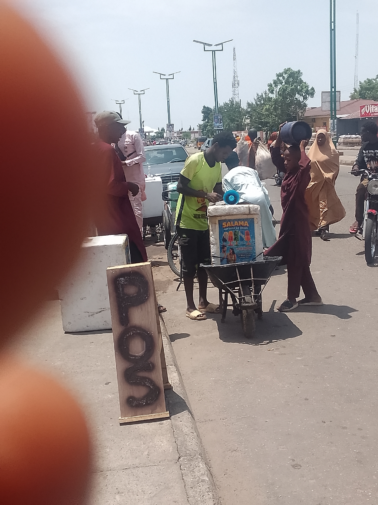 The image depicts an outdoor street scene in an urban area. A man in a yellow shirt stands near a wheelbarrow that holds a cooler labeled "SALAMA POPCICLE ICE CREAM." Another person carries a blue container on their head, near the wheelbarrow. Several people are walking nearby, including a woman in a tan hijab. A sign with the letters "POS" is visible on the left side of the frame. Cars and motorbikes are present on the road. Streetlights and a building with a "Vita" sign are visible in the background. A person's fingers are partly obscuring the left side of the frame.