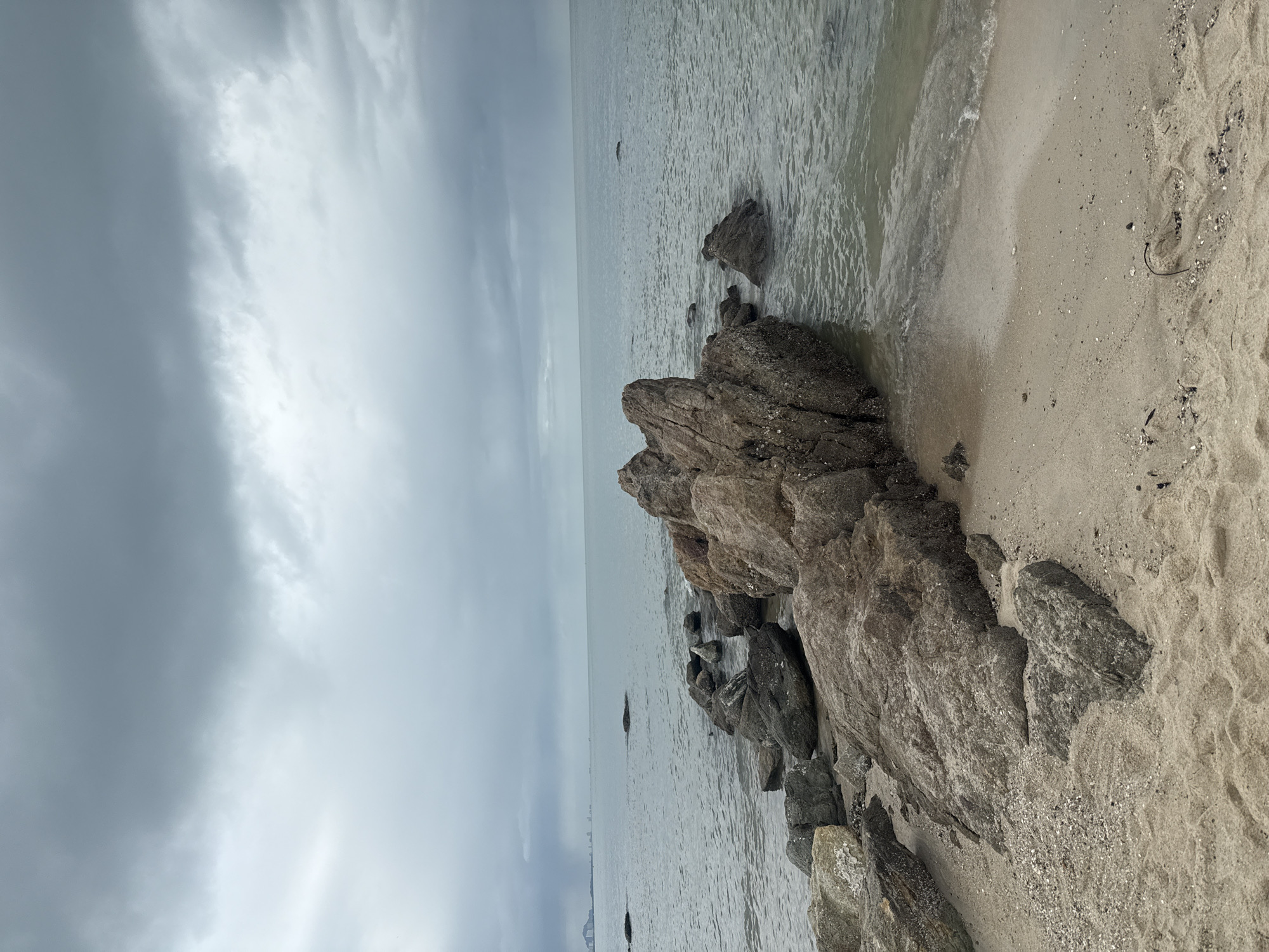 A wide-angle, low-angle shot captures a rocky shoreline at low tide under a cloudy sky. The sand in the foreground is a light tan color, with scattered shells and debris. A cluster of large, dark grey rocks juts out into the calm, grey water. The ocean stretches to the horizon, where faint outlines of buildings and land can be seen under a heavy, overcast sky with visible darker clouds. The scene appears to be at a beach in Hua Hin, Thailand, a popular tourist destination known for its coastline.