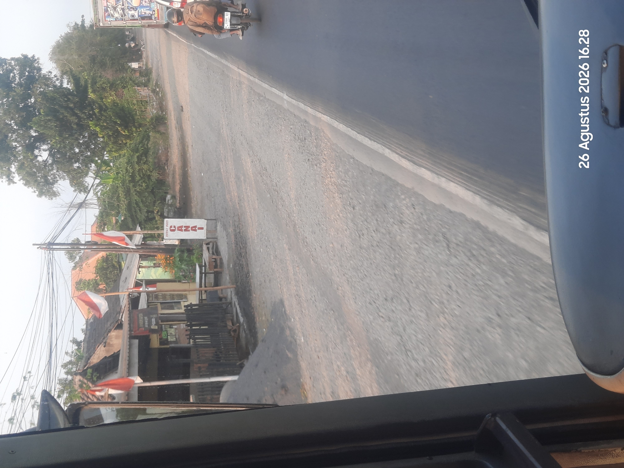 The image depicts a roadside scene in Sukokerto, Indonesia, as indicated by the timestamp "26 Agustus 2026 16.28". In the foreground, a portion of a vehicle's interior is visible on the left, framing a view of the external environment. The roadside features a paved road with a white line, a gravel shoulder, and a small building with Indonesian flags displayed outside. The building appears to be a shop or cafe, with signage that includes the word "CANAI". To the right of the road, a motorcycle with two riders is traveling, one with a backpack. In the background, there are trees and more buildings. The lighting suggests it is late afternoon.