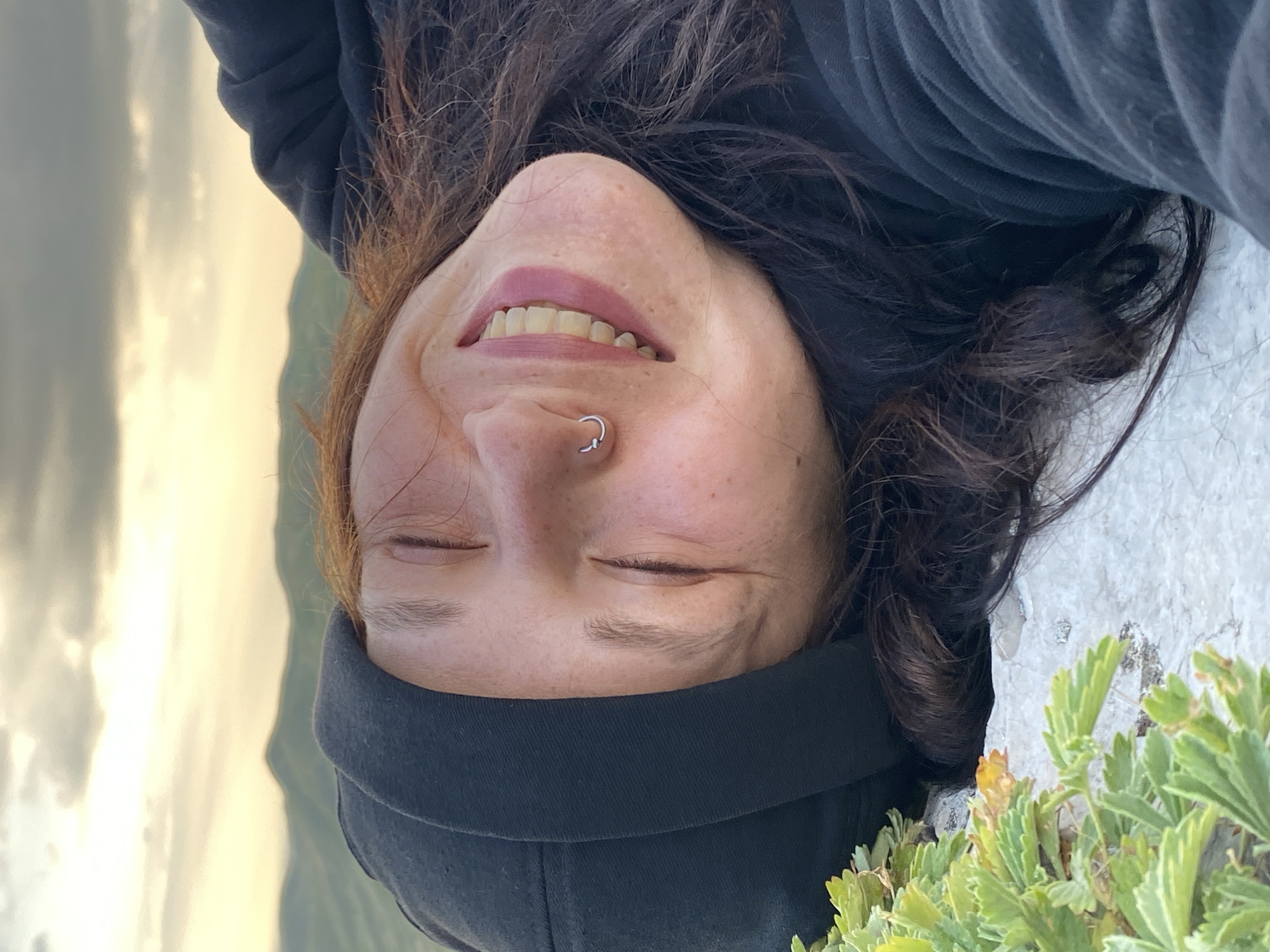A close-up, low-angle, upside-down shot shows a young woman with her eyes closed, smiling broadly, lying on a rock with green plants in the foreground. She is wearing a black beanie and a black hooded sweatshirt. Her reddish-brown hair is spread out around her. A silver hoop nose ring is visible. In the background, rolling hills are visible under a cloudy sky with soft, warm light. The location is stated as Абзаново, Russia.