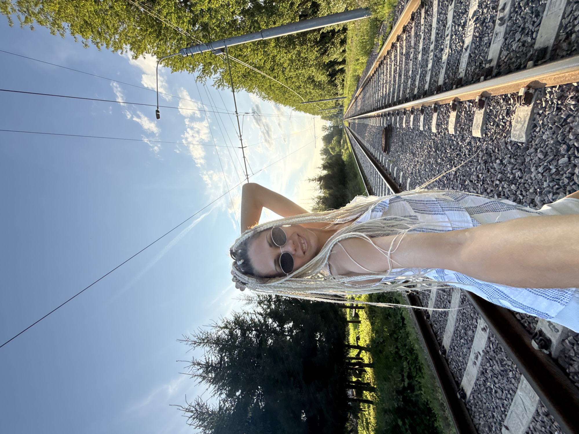 A woman with blonde braided hair and sunglasses takes a selfie on a railway track. She is wearing a white and blue striped dress. The railway track stretches into the distance, flanked by trees on either side. Overhead, power lines and a lamppost are visible against a blue sky with scattered clouds. The scene appears to be in Rusanovo, Russia, during daylight.