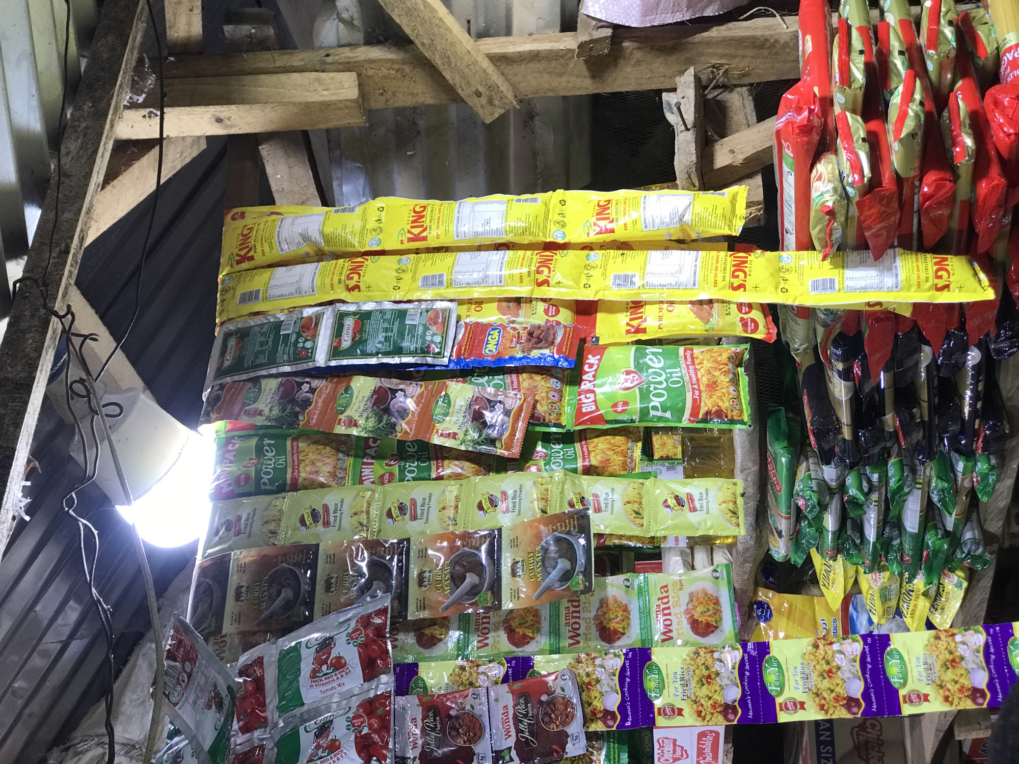 This image captures a close-up view of a market stall or small shop in Abuja, Nigeria, displaying a variety of individually packaged food items. The setting appears rustic, with exposed wooden beams and planks forming the structure of the stall, and corrugated metal sheets visible in the background and to the left. A bright, unshielded artificial light source, likely a bulb, illuminates the scene from the left, suggesting an indoor environment or nighttime operation.
The stall is densely packed with products, primarily small sachets and packets of cooking ingredients and seasonings. Prominently displayed at the top are rows of yellow packets of "DEVON KING'S PURE VEGETABLE OIL." Below these, a diverse range of items includes green and yellow "Power Oil" sachets, some marked with "MINIPAC N200," indicating the price in Nigerian Naira. Other visible products include "Gino" tomato mix, "ONGA Stew" seasoning, "Mr Chef Fried Rice Seasoning Powder," "TIGER CURRY MASALA," "Wonda Jollof Rice Spice," and "Fung Luc Fried Rice Seasonings." Many packages highlight features like "For A Healthy Family," "Mama's Cooking Secret," or "Nutritional Information." On the right, bundles of red and green packaged goods hang vertically, possibly more oil or seasonings. The overall impression is a bustling display of essential cooking ingredients for a local market.