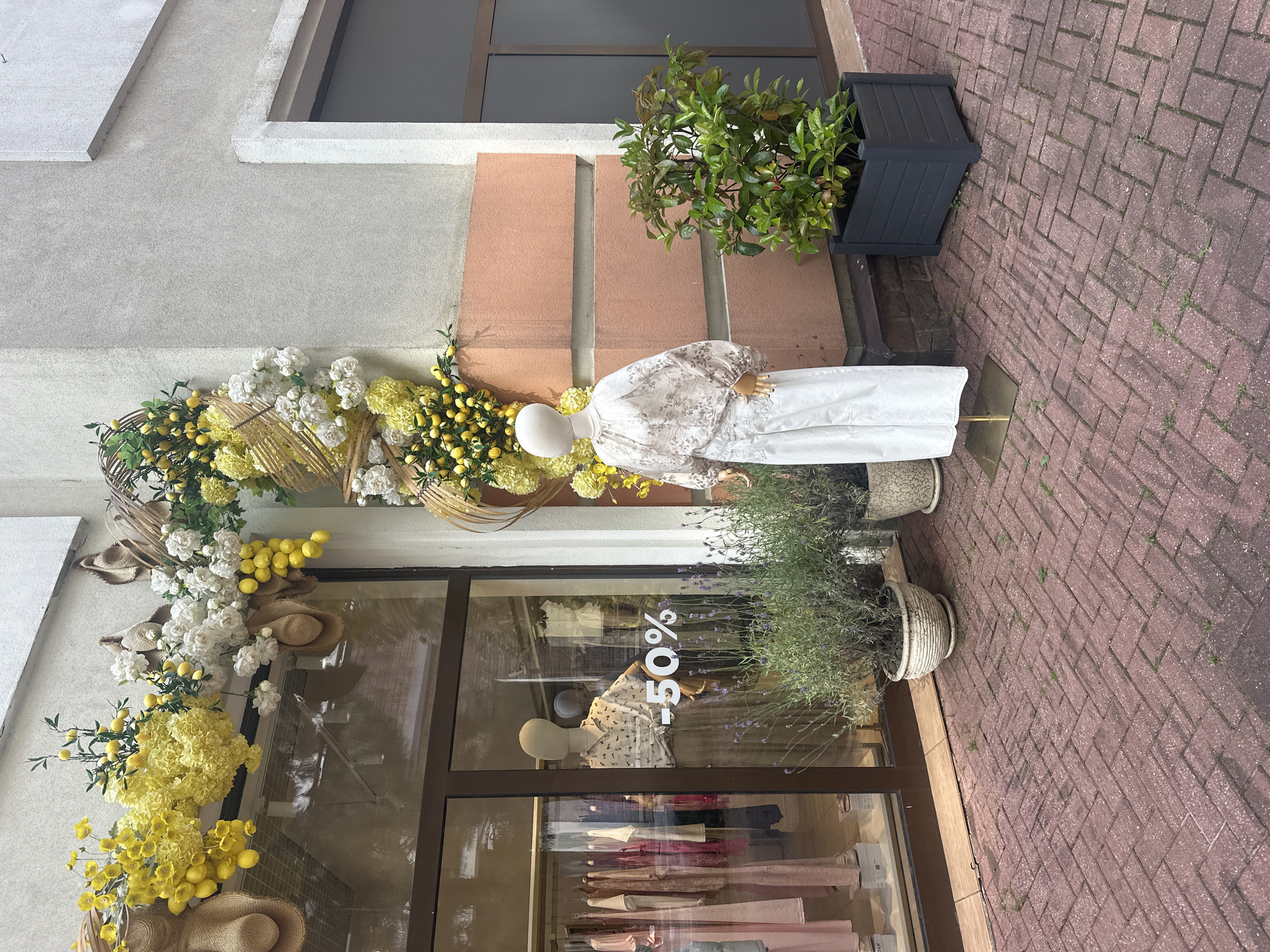 This is a forensic, matter-of-fact description of a commercial storefront in Estosadok, Russia. The scene depicts the exterior of a clothing store with a prominent display in front.
**Key Elements:**
* **Storefront:** The store features a large glass window displaying clothing on racks and a mannequin. A "50%" sale sign is visible on the glass. The building facade is characterized by a light-colored upper section and a pale pink lower section with horizontal banding.
* **Mannequins:** Two mannequins are present. One is inside the store, facing the window and wearing a patterned shirt and skirt. Another is outside the store, positioned to the right of the entrance, dressed in a white blouse with voluminous sleeves and white trousers.
* **Floral and Decorative Elements:** An elaborate floral arrangement featuring yellow flowers, lemons, and straw hats is draped above the store window and extends around the corner.
* **Potted Plants:** Two potted plants are situated on the pavement in front of the store. The plant on the left is a lavender bush in a decorative white pot. The plant on the right is a green leafy shrub in a dark gray rectangular planter.
* **Pavement:** The ground in front of the store is paved with reddish-brown brick laid in a herringbone pattern. Small weeds are visible growing between some of the bricks.
* **Environment:** The scene appears to be an outdoor sidewalk or pedestrian area in front of a commercial establishment. The lighting suggests daytime.
**Absence of Information:**
* No specific details about the clothing store's name or brand are discernible.
* No other people are clearly visible or engaged in any discernible actions within the frame.
* The broader urban context beyond the immediate storefront is not evident.