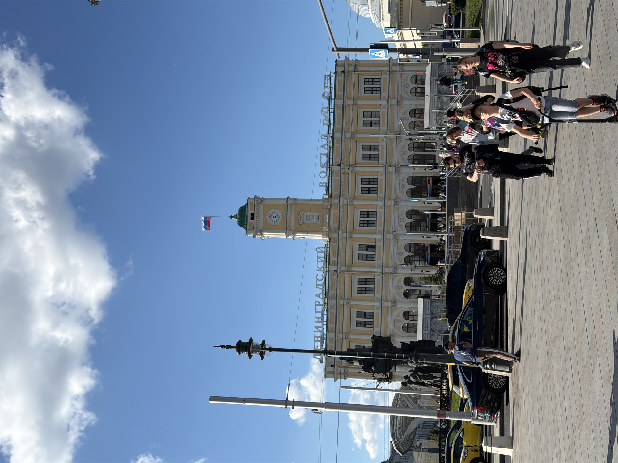 The image captures an outdoor urban scene in Moscow, Russia, dominated by the historic Leningradsky Station under a clear, bright blue sky with some scattered white clouds. The photograph is taken during daytime, evidenced by the strong natural light and visible shadows.
The most prominent feature is the large, multi-story yellow building of Leningradsky Station, which features classical architectural elements, numerous windows, and white decorative details. A tall clock tower, topped with a green dome and a spire from which a Russian flag proudly flies, rises from the left side of the station building. Above the main entrance or a central section of the building, large white letters spell out "ВОКЗАЛ ЛЕНИНГРАДСКИЙ" (Vokzal Leningradskiy), which translates to "Leningradsky Station".
In the foreground and midground, a wide paved plaza or street stretches out. Several people are visible across this area. On the right, a group of young individuals, appearing to be teenagers or young adults, are standing and looking in various directions; one person is next to a scooter. Further back on the right, several cars, including yellow taxis and darker vehicles, are parked along the edge of the paved area. A man in a blue shirt is standing near one of these parked cars. Various streetlights and poles are distributed throughout the scene, along with blue and white road signs. The overall atmosphere suggests a busy but ordinary day at a major transport hub.