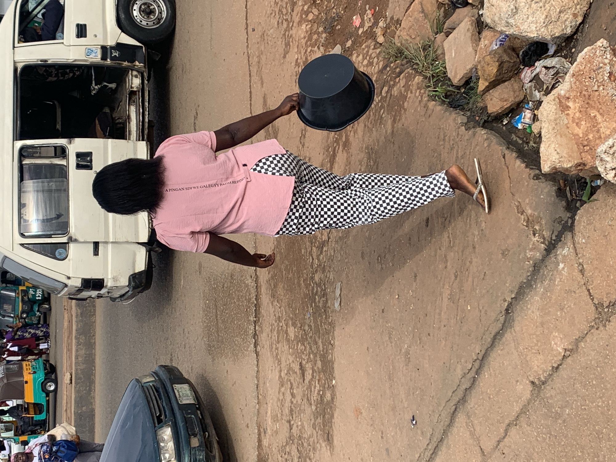 An African woman walks away from the camera, carrying a black bucket in her right hand, on a dirt and asphalt road in Gura Top, Nigeria. She wears a pink t-shirt and black and white checkered pants. She is walking along a cracked concrete sidewalk that is partially covered in dirt and debris. To the right of the sidewalk is a pile of large rocks and trash. In the background, a white van with its doors open is parked on the road, and further in the background, there are yellow and green auto-rickshaws. The scene depicts a typical street in Nigeria, with a mix of vehicles, people, and unpaved road surfaces.