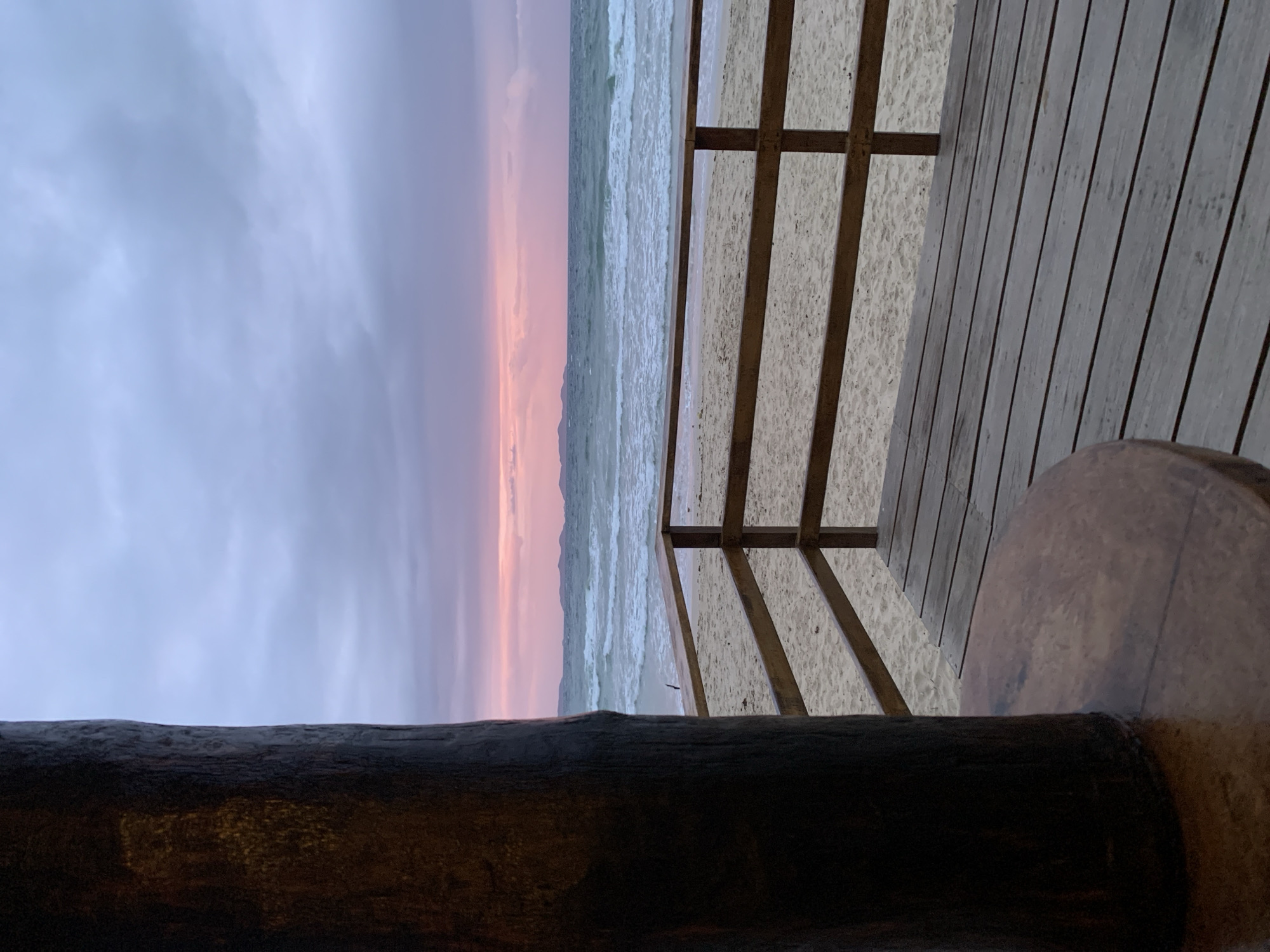 The image depicts a serene beach scene at dusk, likely in El Tirano, Venezuela, given the context provided. In the foreground, a large, dark, cylindrical wooden post is partially visible on the left, with a circular wooden table in front of it. To the right, a wooden deck with parallel planks leads to a wooden railing that overlooks a sandy beach. Beyond the beach, the ocean stretches to the horizon, with gentle waves breaking on the shore. In the distance, a hazy outline of islands or a coastline can be seen. The sky is filled with dramatic clouds, painted with hues of purple and pink from the setting sun, creating a moody and tranquil atmosphere. No people are present in the frame.