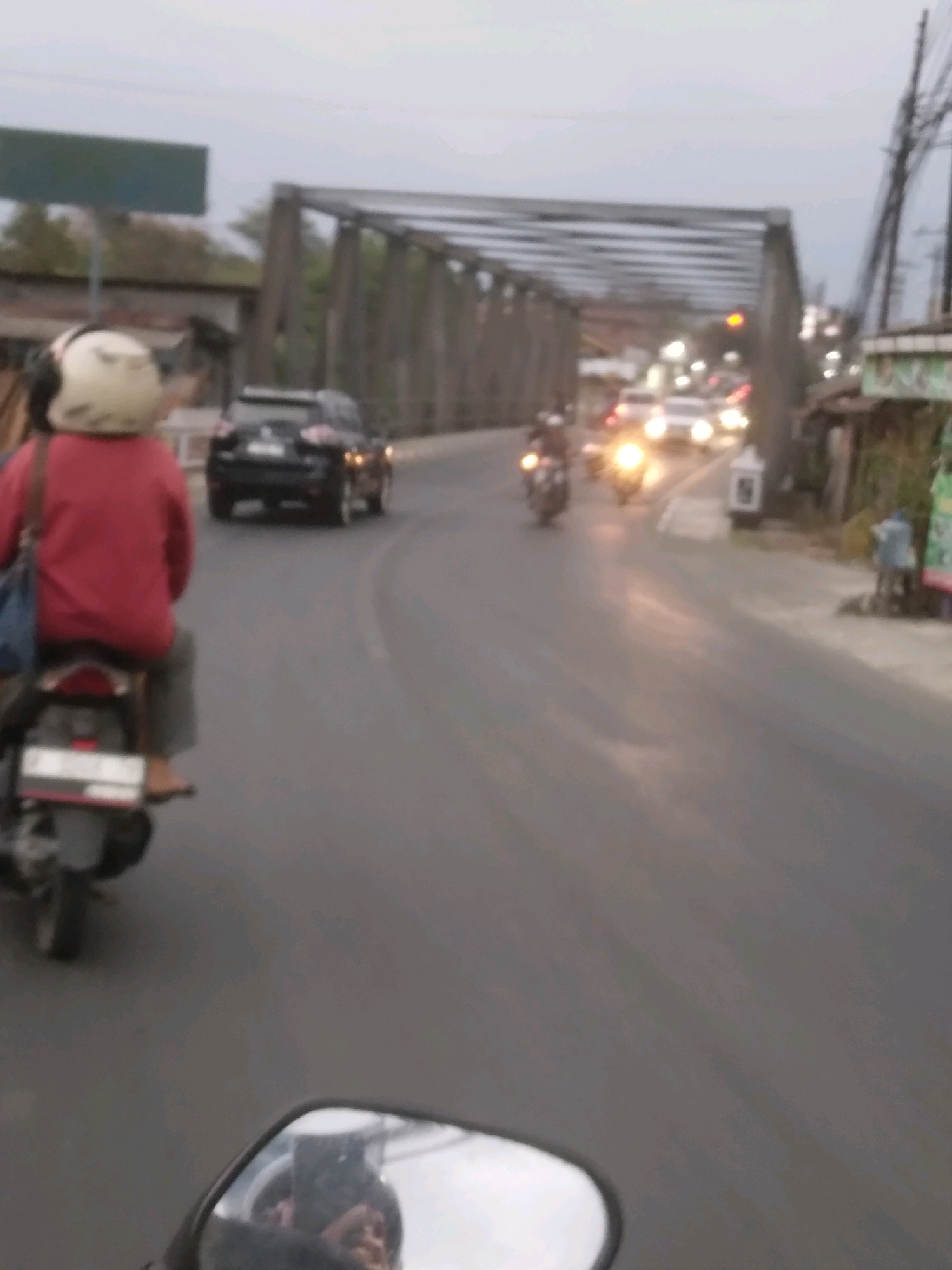 The image captures a scene on a road in Sokaraja, Indonesia, likely during dusk or early morning, given the dim lighting and the headlights of vehicles. A bridge, appearing to be made of steel trusses, dominates the background, spanning across the road. In the foreground, a person is riding on a motorcycle, seen from behind. They are wearing a red jacket and a white helmet. Another motorcycle is visible on the road ahead, with its headlights on. Further down the road, several cars are driving, their headlights also illuminated. The road surface is asphalt, and there are some buildings and trees visible on the sides of the road. The overall impression is one of moderate traffic on a road leading up to or under a bridge in a town or city setting.