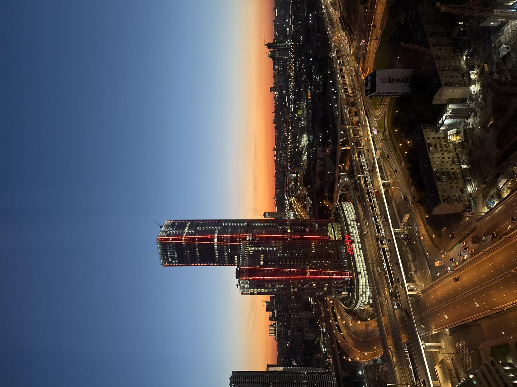 The image is an aerial view of the "ICITY" complex in Khimki, Russia, at dusk. The most prominent structures are two tall, modern skyscrapers illuminated with bright red vertical lights. The taller skyscraper has "ICITY" spelled out in large red letters at its base. The sky transitions from a deep blue at the top to an orange and pink gradient near the horizon, indicating sunset or sunrise.
Below the skyscrapers, a complex network of elevated highways and roads is visible, with cars and trucks moving along them, their headlights and taillights creating streaks of light. The surrounding urban landscape consists of numerous buildings, some appearing to be residential blocks and others commercial structures, all lit up with various interior and exterior lights. Several construction cranes are visible in the foreground, suggesting ongoing development in the area. The overall scene depicts a dynamic urban environment at the transitional time of day.