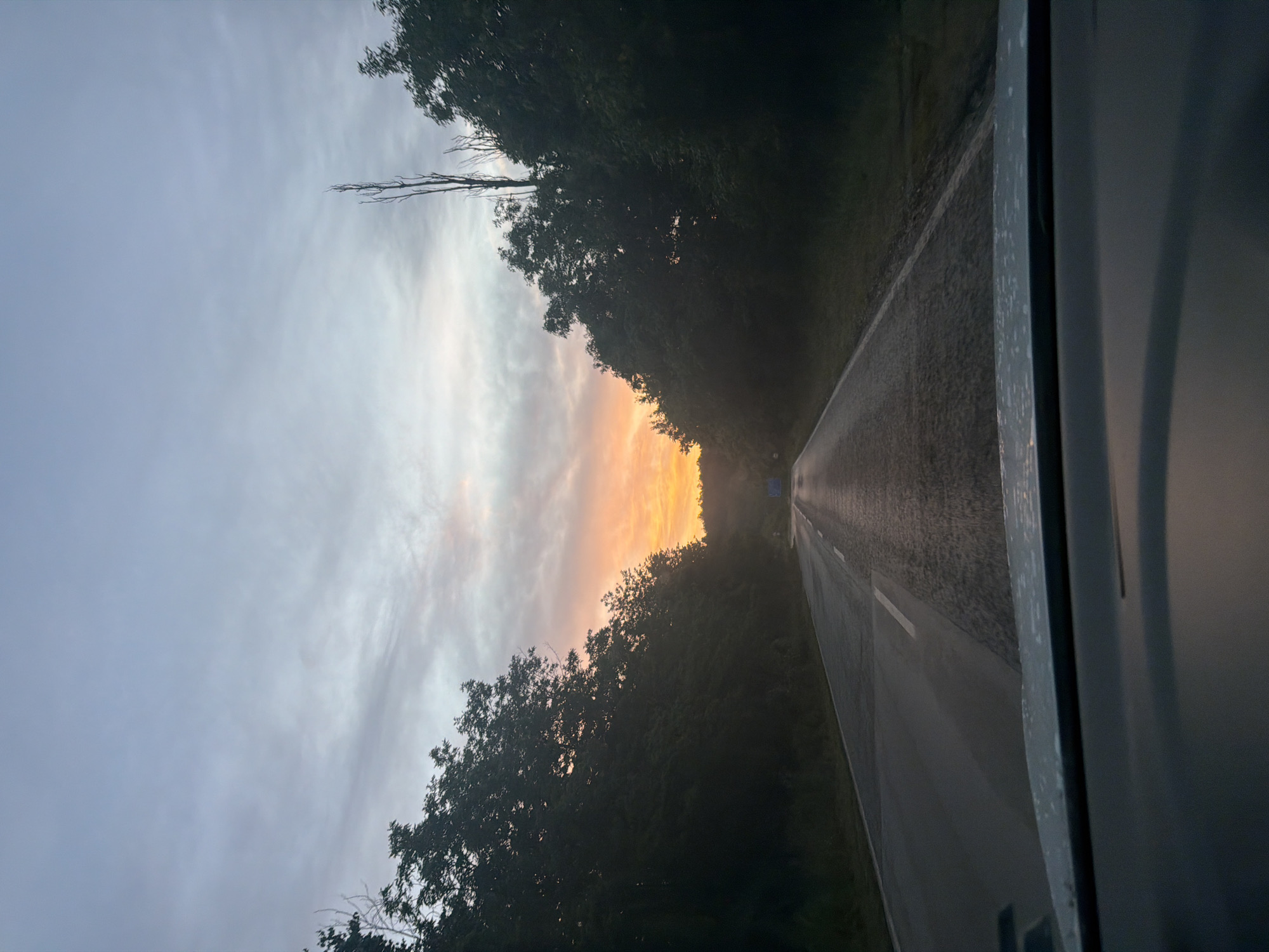 This is a first-person perspective, wide-angle photograph taken from inside a vehicle, looking forward. The scene captures a road with trees lining both sides, leading towards a sunrise. The sky is a gradient of pale blue to grey, with streaks of orange and pink from the rising sun visible in the distance. The road appears wet, possibly from recent rain, and has a white line marking its center. The foreground shows the dashboard and windshield of the vehicle. The environment suggests a rural or semi-rural setting, possibly on the outskirts of the city of Хаджийско, Bulgaria. There are no people or animals visible in the frame. The overall mood is serene and contemplative, indicative of an early morning drive.