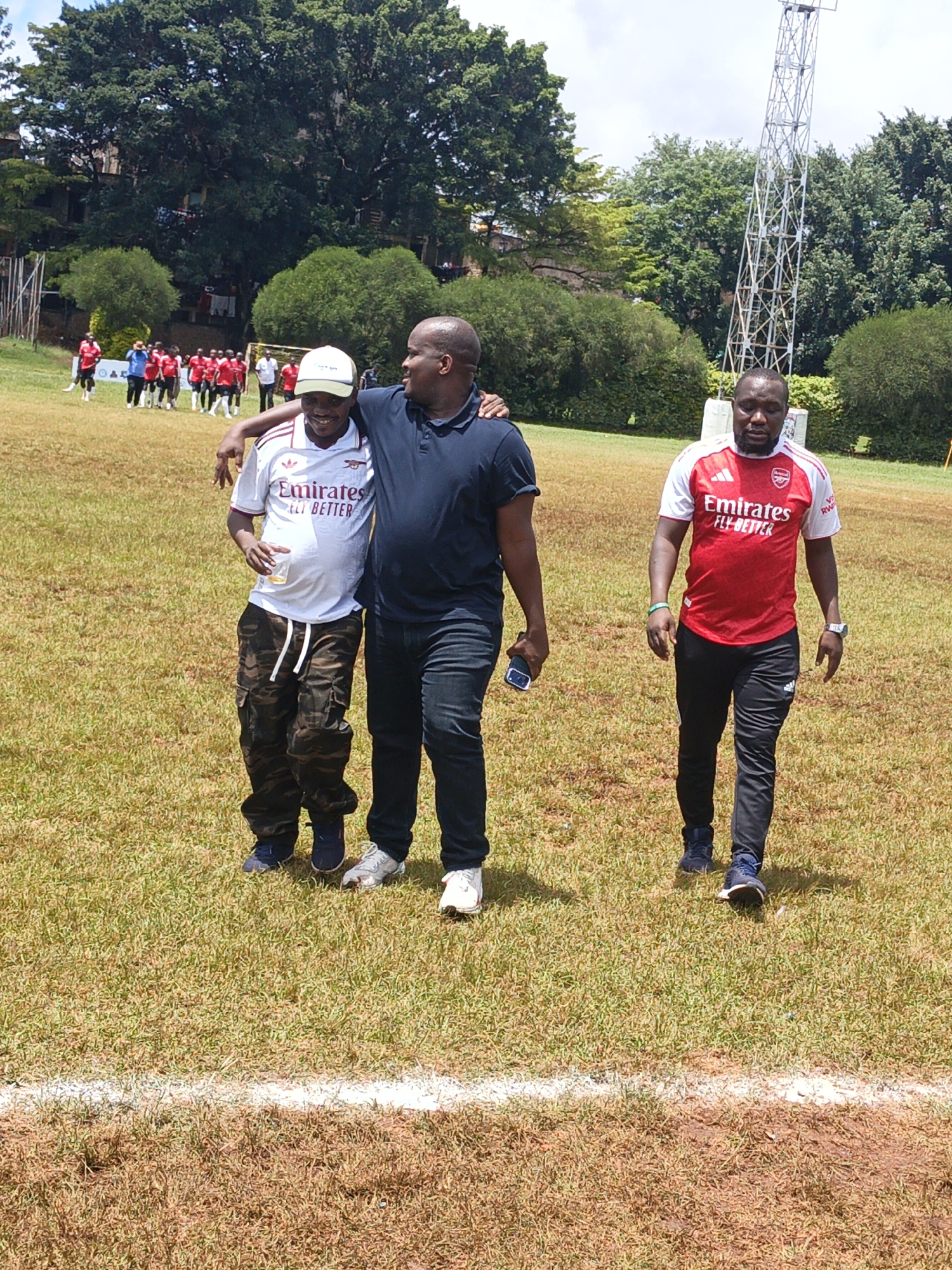 The image depicts a grassy field, possibly a sports field, in Nairobi, Kenya. In the background, a group of people wearing similar red and white uniforms are visible near a soccer goal. A tall, lattice communication tower stands in the background on the right side of the frame.
In the foreground, three men are walking across the field. The man on the left wears a white sports jersey with the words "Emirates FLY BETTER", camouflage pants, and a baseball cap. He has a drink in his hand. The man in the center has his arm around the man on the left. He wears a navy blue polo shirt and dark jeans. The man on the right wears a red and white sports jersey with the same "Emirates FLY BETTER" branding and black sweatpants. All three men are facing towards the viewer. A white line runs horizontally across the foreground, indicating the boundary of the field.