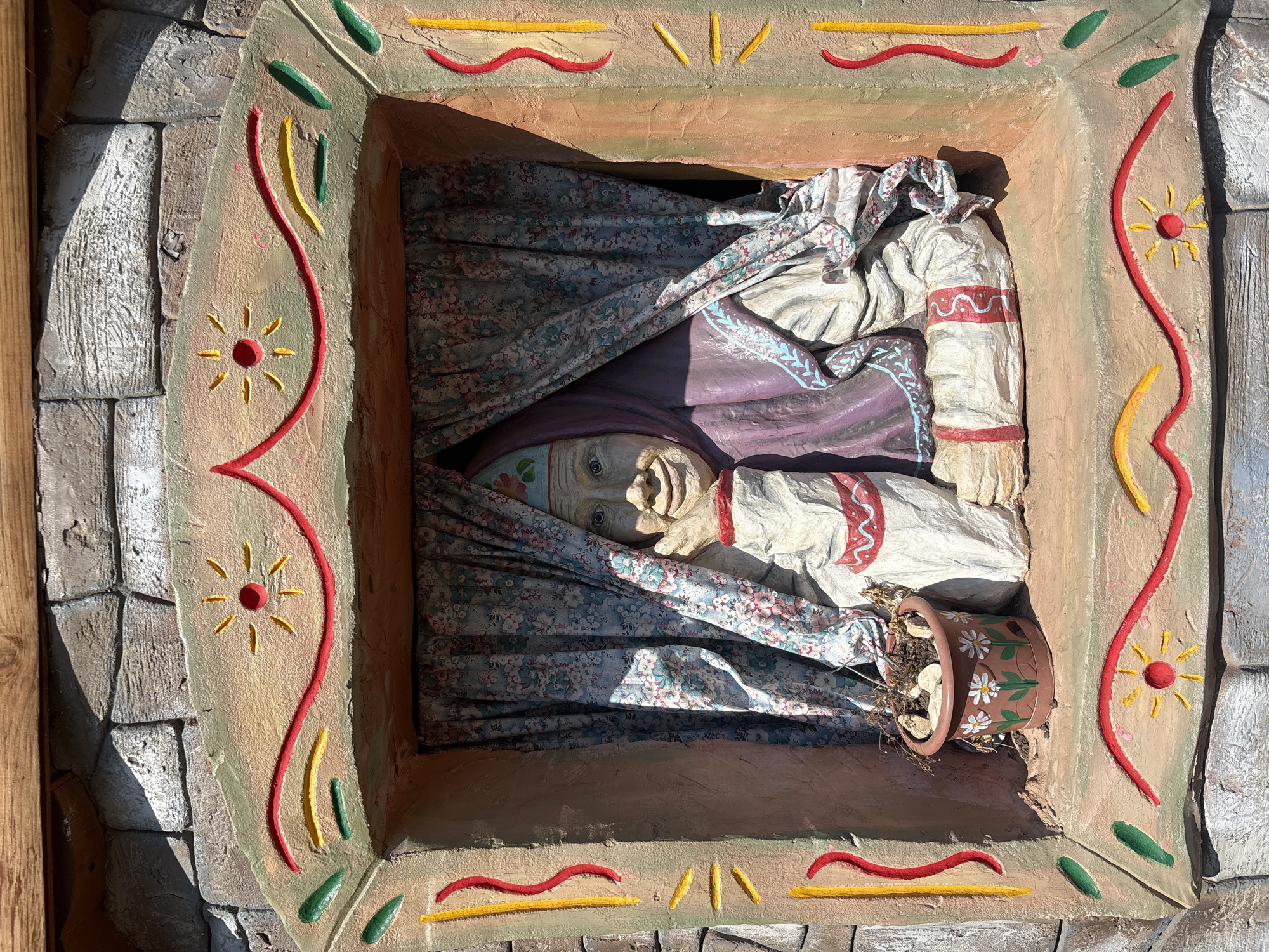 The image shows a three-dimensional art installation located in the city of Chaikovsky, Russia. The installation depicts an elderly woman, seemingly in a coffin or bed-like structure, adorned with fabric resembling bedding. The woman wears a head covering, and her hands are positioned on her chest. A flower pot with white daisy-like decorations is located near her head or the edge of the "coffin." The structure surrounding the woman is rectangular and decorated with painted details, including stylized suns, curved lines in yellow and red, and green shapes. The outer layer of the structure resembles stone or brickwork.
