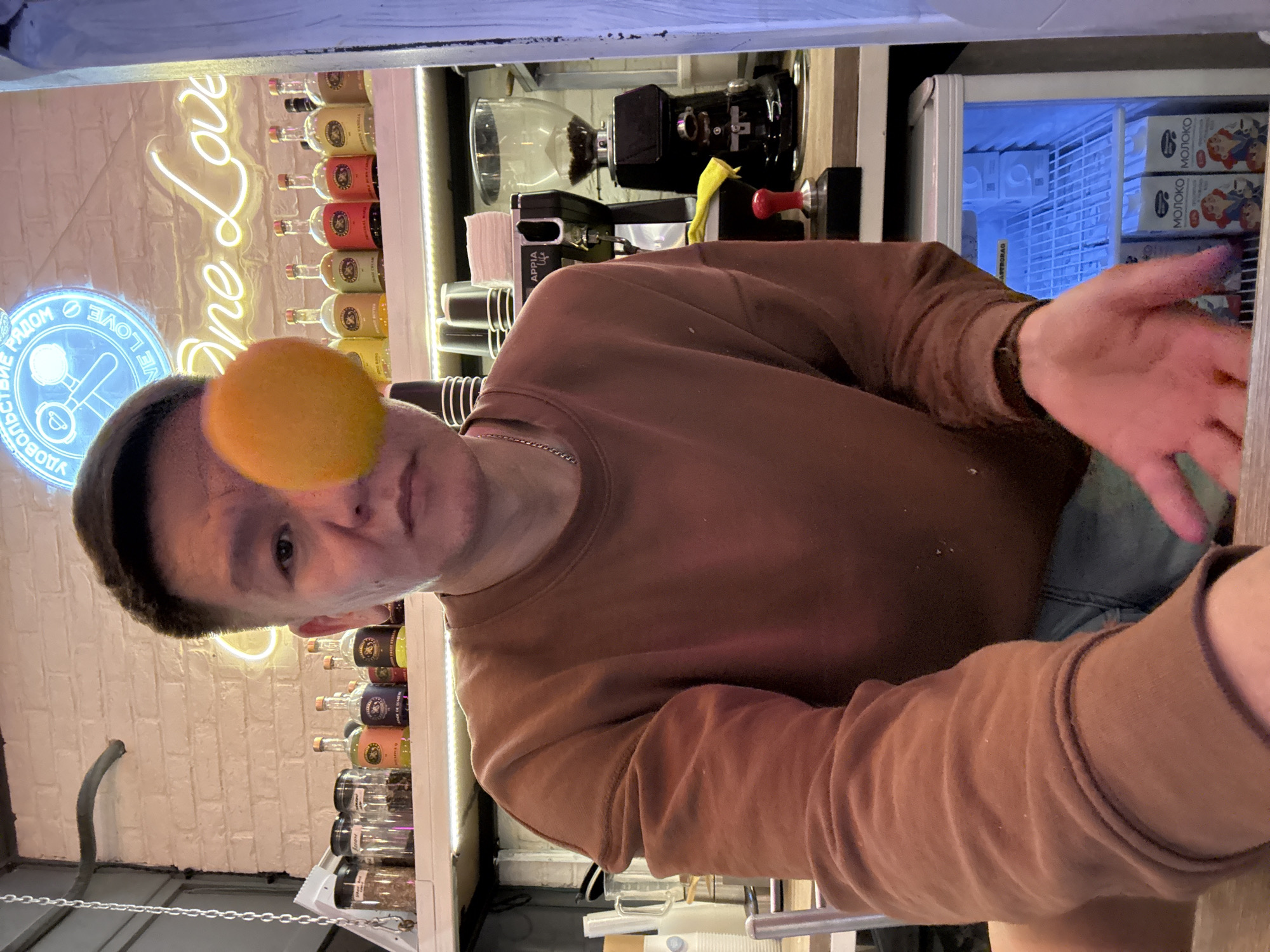 A man is taking a selfie in a brightly lit establishment. He is wearing a brown crew neck sweatshirt and denim shorts. An orange is positioned in front of his right eye. Behind him, shelves are stocked with various bottles and jars. A neon sign that reads "One Love" is illuminated on the wall. A coffee grinder and a small refrigerator are also visible. The overall environment suggests a cafe or bar.