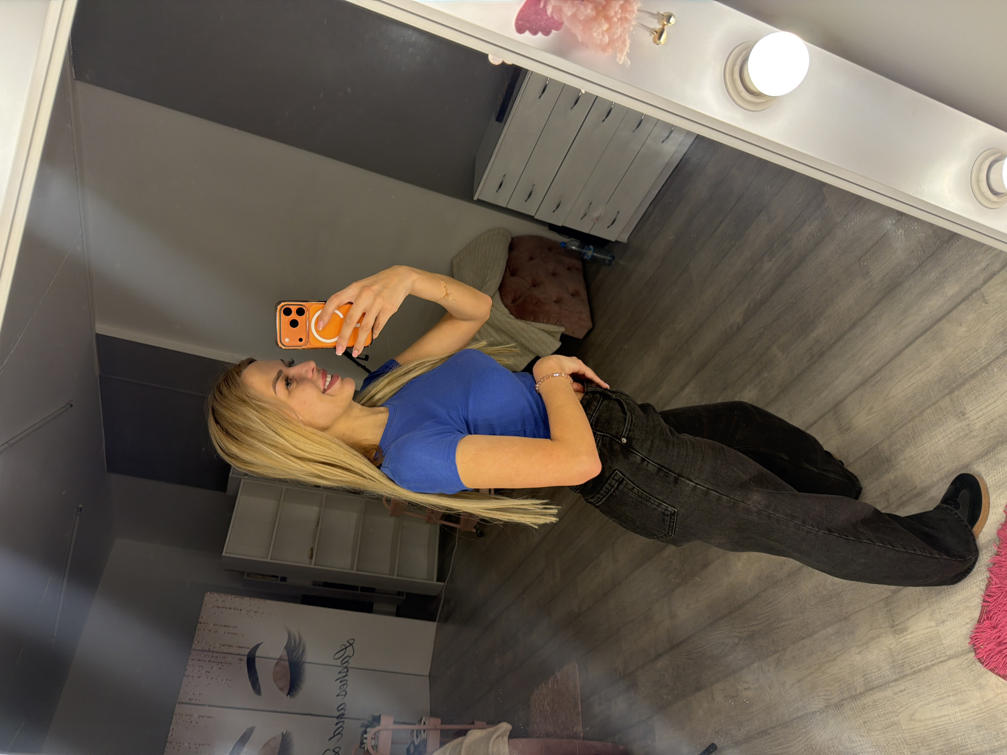 A young woman with blonde hair is taking a mirror selfie in what appears to be a dressing room or vanity area. She is wearing a blue t-shirt and dark wash jeans. She is holding an orange smartphone in her right hand, angled to capture her reflection. She is smiling and looking at her reflection.
In the background, visible in the mirror, is a vanity mirror with lights, a grey wall, and a wooden floor. There is also a white dresser with drawers, a pink tufted ottoman or stool, and a framed picture on the wall featuring eyelashes. The overall impression is of a personal space, possibly for getting ready. The lighting suggests it could be daytime or well-lit artificial lighting. There is no visible text in the image.