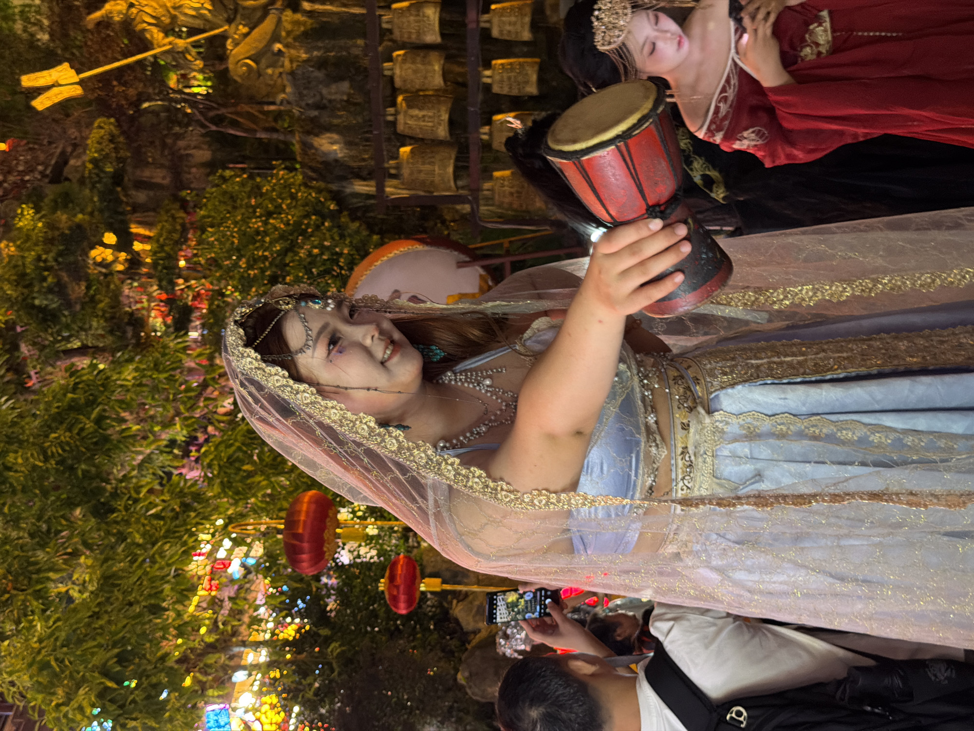 In Yuzhong District, China, a woman adorned in a light blue, traditional Chinese-style outfit with gold embellishments and a sheer veil over her head is performing. She is holding a small red and black drum in her right hand, poised to strike it. Her head is tilted slightly, and she is smiling as she looks toward her right. In the background, to the right of the performing woman, another woman in a red traditional Chinese outfit is looking down at her phone. Behind them, a wall of golden bells is visible, and further back, a statue of a figure holding a spear stands amidst lush greenery adorned with colorful lights and red lanterns. To the left of the performing woman, a young person with short dark hair is holding up a smartphone to capture the scene. The overall environment appears to be an outdoor night event, possibly a festival or cultural performance, illuminated by artificial lighting.