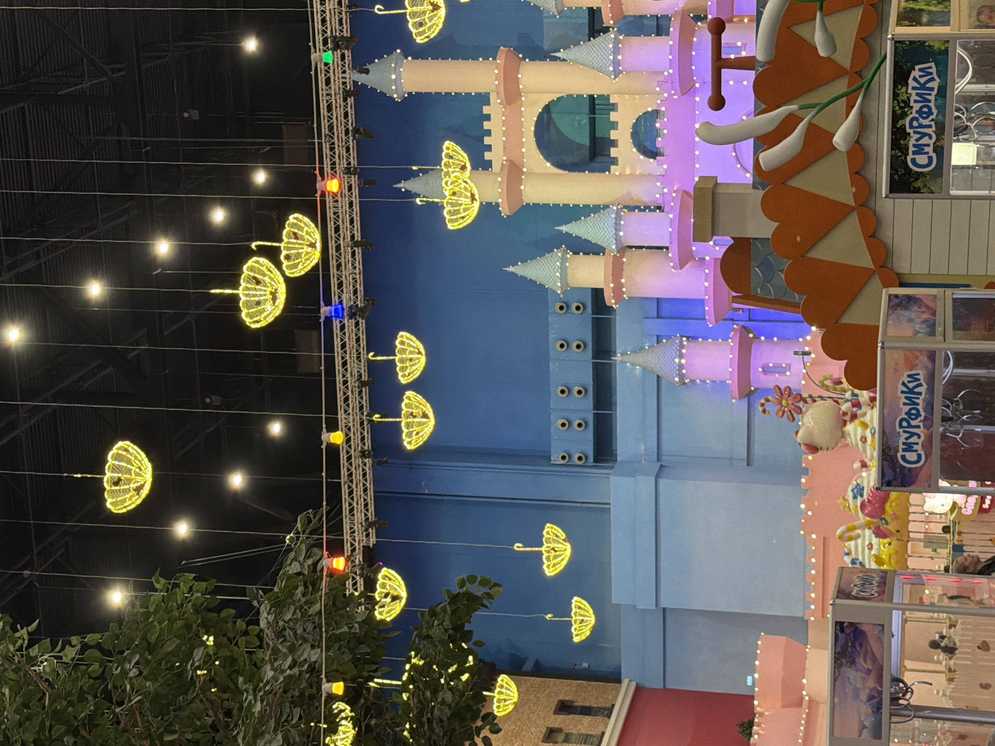 The image captures an indoor amusement park in Vidnoye, Russia, likely within the Smurfs themed area. Suspended from the ceiling are numerous decorative yellow lights shaped like open umbrellas, illuminating the scene. In the background, a large, multi-turreted castle structure painted in pastel pink and yellow with white lights is a central focal point. In the foreground, several arcade machines are visible, one with "СМУРФИКИ" (Smurfs) signage, and another displaying a colorful image of what appears to be a fantasy landscape. A large artificial tree with green leaves and yellow lights is positioned on the left side of the frame. Several people are present, including individuals operating or observing the arcade machines. The environment is brightly lit and decorated with a whimsical, child-friendly theme.