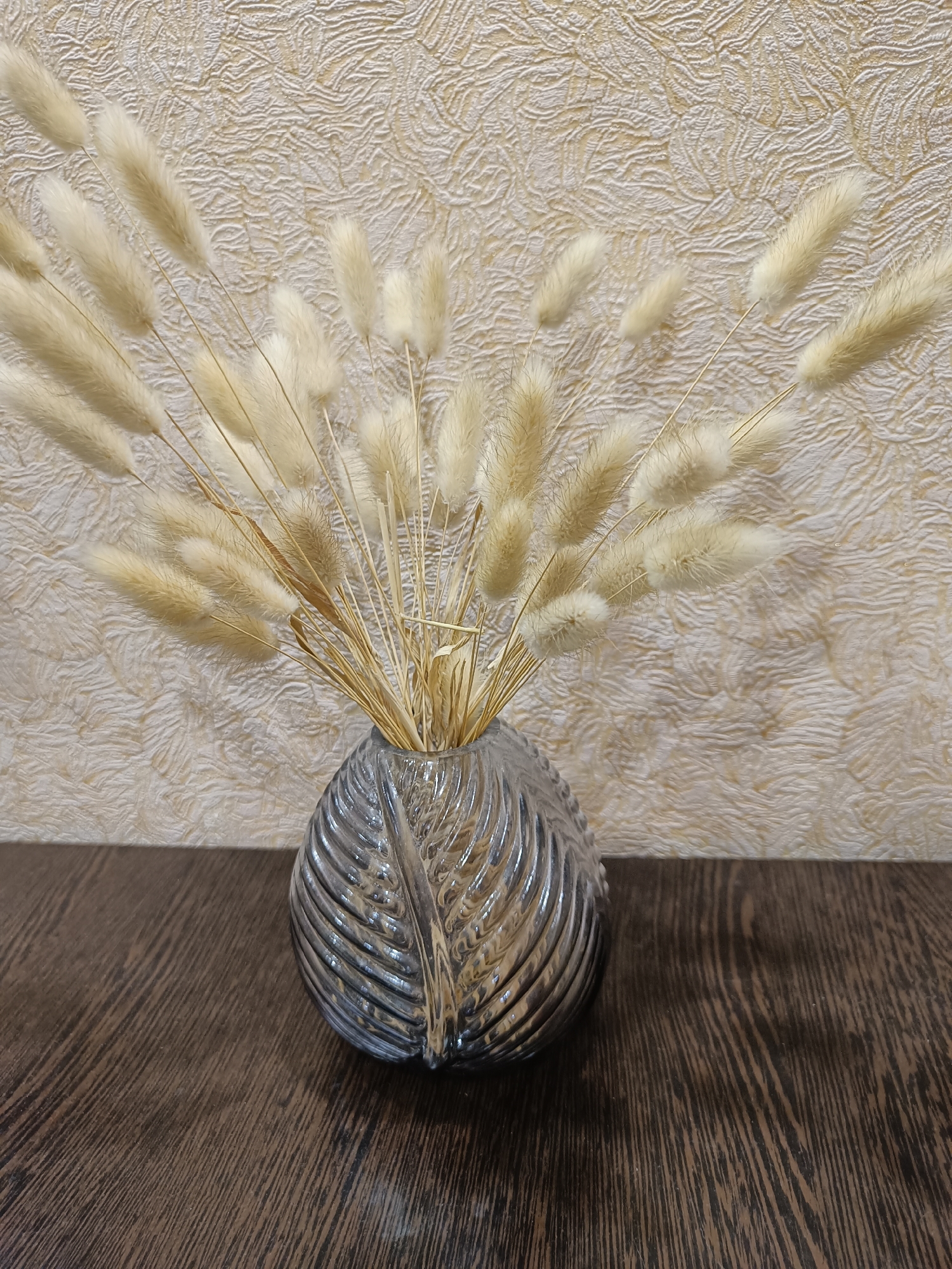 A close-up, eye-level shot features a translucent, fluted glass vase with a ribbed, leaf-like pattern. The vase is filled with a dried arrangement of beige bunny tail grasses, their fluffy tips fanned out against a textured, cream-colored wall. The vase rests on a dark brown wooden surface, its grain visible in diagonal lines. The scene is indoors, and there are no people or animals present. The lighting is soft, creating subtle shadows. The image does not provide any information that can be used to determine its location in the city of Vichuga, Russia.