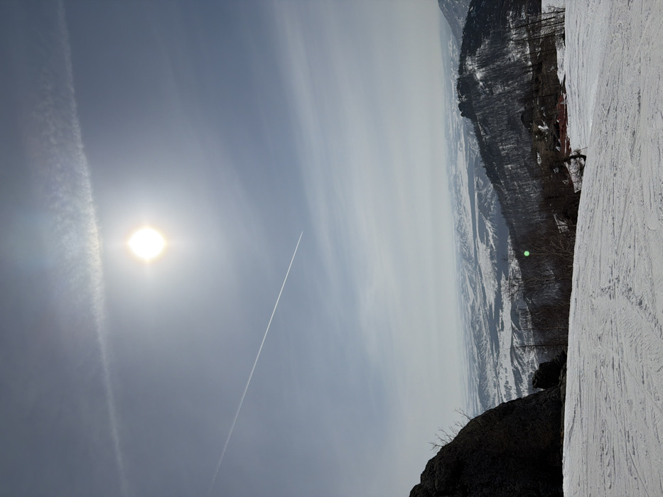 The image depicts a panoramic winter landscape from a snow-covered mountain slope in Zelenaya Polyana, Russia. The sky, occupying the upper two-thirds of the frame, is light blue with a brilliant, circular sun positioned in the upper center, generating a prominent lens flare and illuminating the scene directly. A linear contrail from an aircraft extends diagonally across the sky from the upper left to the lower-mid left. The foreground features a wide expanse of disturbed snow, indicating a ski slope with multiple ski tracks. To the far left, a large, dark rock formation rises, partially framing the view. The midground showcases snow-dusted mountains and hills sparsely covered with bare deciduous trees and some evergreen foliage. A cluster of small, red-roofed buildings is discernible on a distant slope to the right. A distinct green lens flare artifact is present in the lower-midground. The background comprises an expansive, hazy vista of snow-covered plains and rolling terrain stretching to the horizon under a pale sky. No individuals are visible.