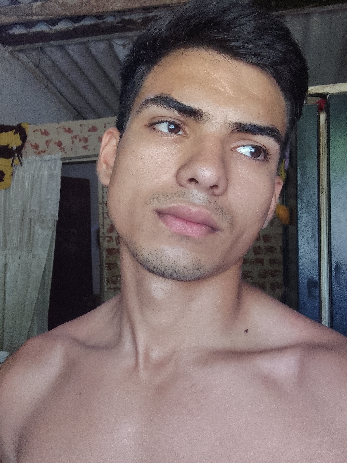 The image displays a close-up portrait of a young adult male in an indoor setting within Juan Griego, Venezuela. The subject, bare-chested and facing generally forward, has a medium complexion, dark short hair, and dark eyes looking towards the upper right. A healed linear scar is visible on his forehead above his left eyebrow. Light stubble covers his chin and jawline. A small, dark mole or blemish is present on his right collarbone. The background features an interior room with visible structural elements. To the left, a light-colored, possibly sheer curtain hangs, partially obscuring a patterned wall and a yellow and dark-striped fabric or blanket. To the right, a dark vertical frame or door is partially visible, with a faint brick-patterned wall beyond it and a small yellow object near the top. The ceiling area reveals exposed, corrugated metal roofing with supporting wooden beams. The lighting appears to be ambient, illuminating the subject from the front or right. The male is not interacting with other individuals and appears to be posing for the photograph.
