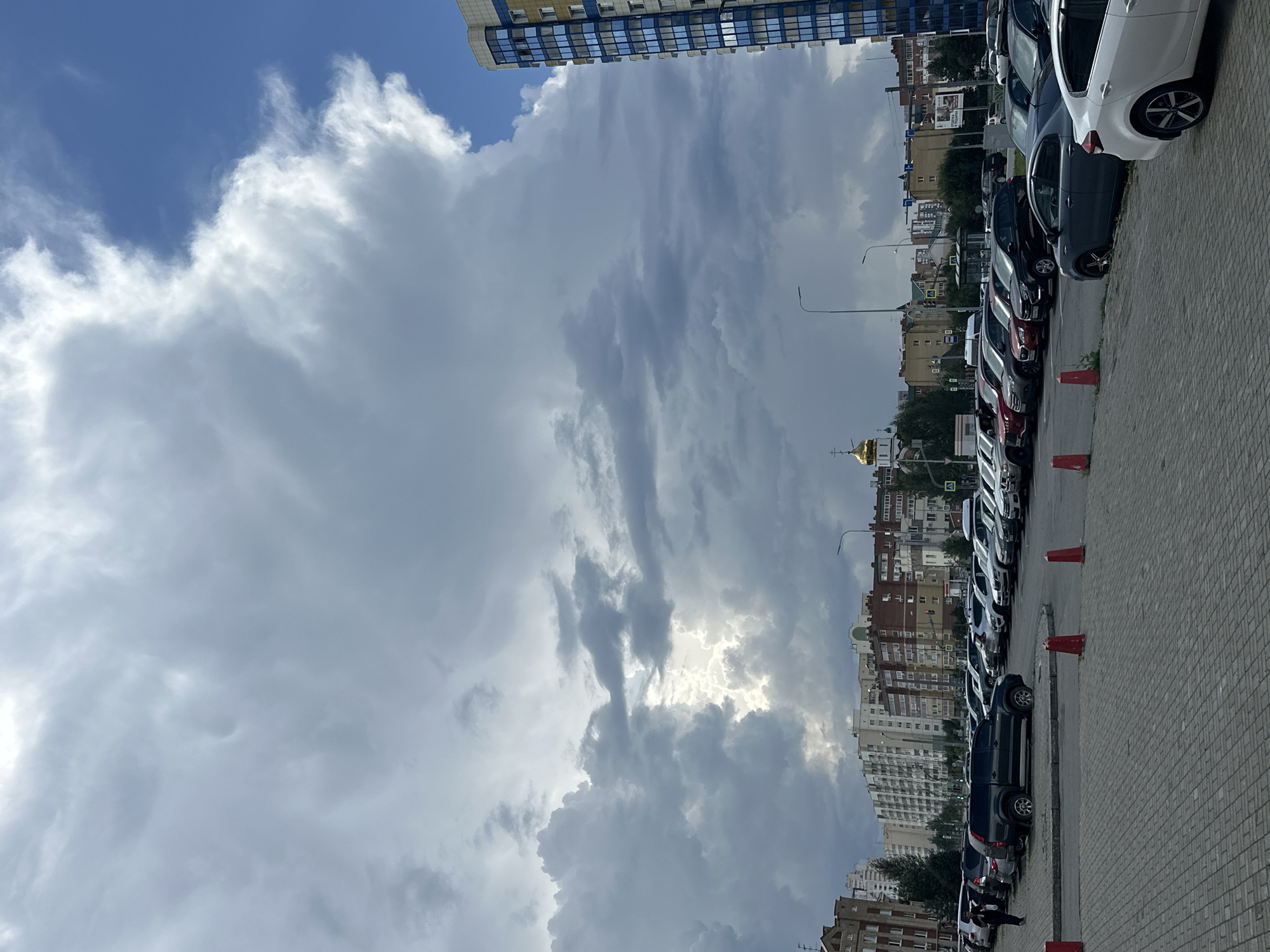 The image depicts a wide-angle view of a parking lot in Yekaterinburg, Russia, under a dramatic, cloudy sky with patches of bright blue. In the foreground, a paved parking area is visible, with red traffic cones marking a section. Numerous cars are parked in rows, including SUVs, sedans, and hatchbacks. A dark SUV is prominently positioned in the middle ground, with a person walking nearby.
Beyond the parking lot, a cityscape unfolds. Buildings of varying heights and architectural styles line the horizon, featuring brick and modern apartment complexes. A distinctive golden-domed Orthodox church is visible amongst the buildings, a common sight in Russian cities. To the right, a tall, modern residential building with a blue and yellow facade stands out. Streetlights and trees are scattered throughout the scene. The overall impression is of an urban environment on a day with dynamic weather patterns.