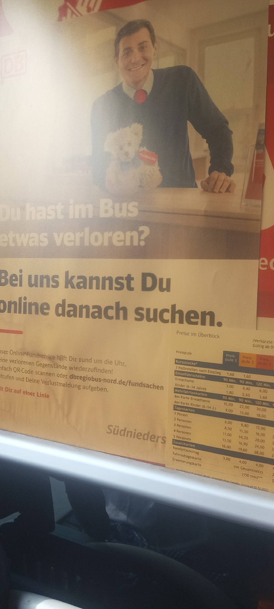 The image shows a close-up of a printed advertisement, likely inside a bus. The advertisement is for DB (Deutsche Bahn) and promotes their online lost and found service. The text reads "Du hast im Bus etwas verloren? Bei uns kannst Du online danach suchen" ("Did you lose something on the bus? With us you can search for it online"). The advertisement also includes a photo of a smiling man and a small white teddy bear, with the text "Fundsache" (Lost property) on it. Below the main text, there's information about fares ("Preise im Überblick"), listing prices for different ticket types. The location in Jos, Nigeria is not specified and this appears to be an advert for German services.