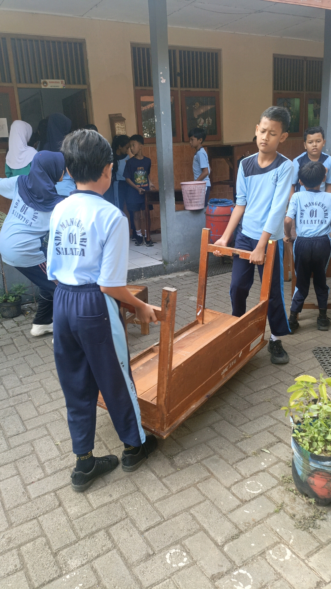 In Salatiga, Indonesia, two boys are working together to move a wooden desk on a paved outdoor walkway. The boy on the left, facing away from the camera, wears a blue shirt with "SDN MANGUNSARI 01 SALATIGA" printed on the back and dark blue pants with a light blue stripe. He is holding the desk by its side. The boy on the right, facing towards the camera, is wearing a similar blue shirt and dark blue pants. He is holding the desk by the legs, supporting his partner. The desk is made of light-colored wood and appears to be flipped upside down. The background shows a building with windows and a pillar. Other children and adults are visible in the background, some observing, others engaged in their own activities. The ground is paved with rectangular bricks. A potted plant sits to the right of the frame.