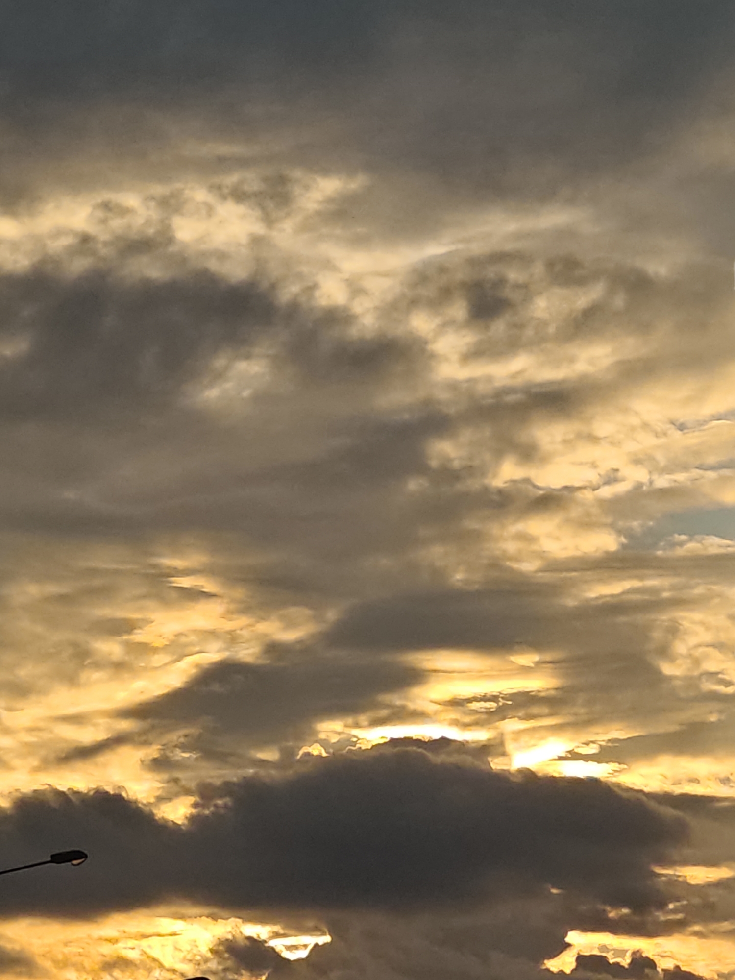 The image is a close-up, upward-facing shot of a cloudy sky at dusk. The clouds are dark gray and heavy, with streaks of bright yellow and orange light filtering through them, indicating the sun is low on the horizon. A streetlamp with a dark, curved pole extends from the lower left corner of the frame, silhouetted against the sky. The overall impression is dramatic and atmospheric, with the light and shadow creating a sense of depth and texture within the clouds. The location is identified as Shcherbinka, Russia.
