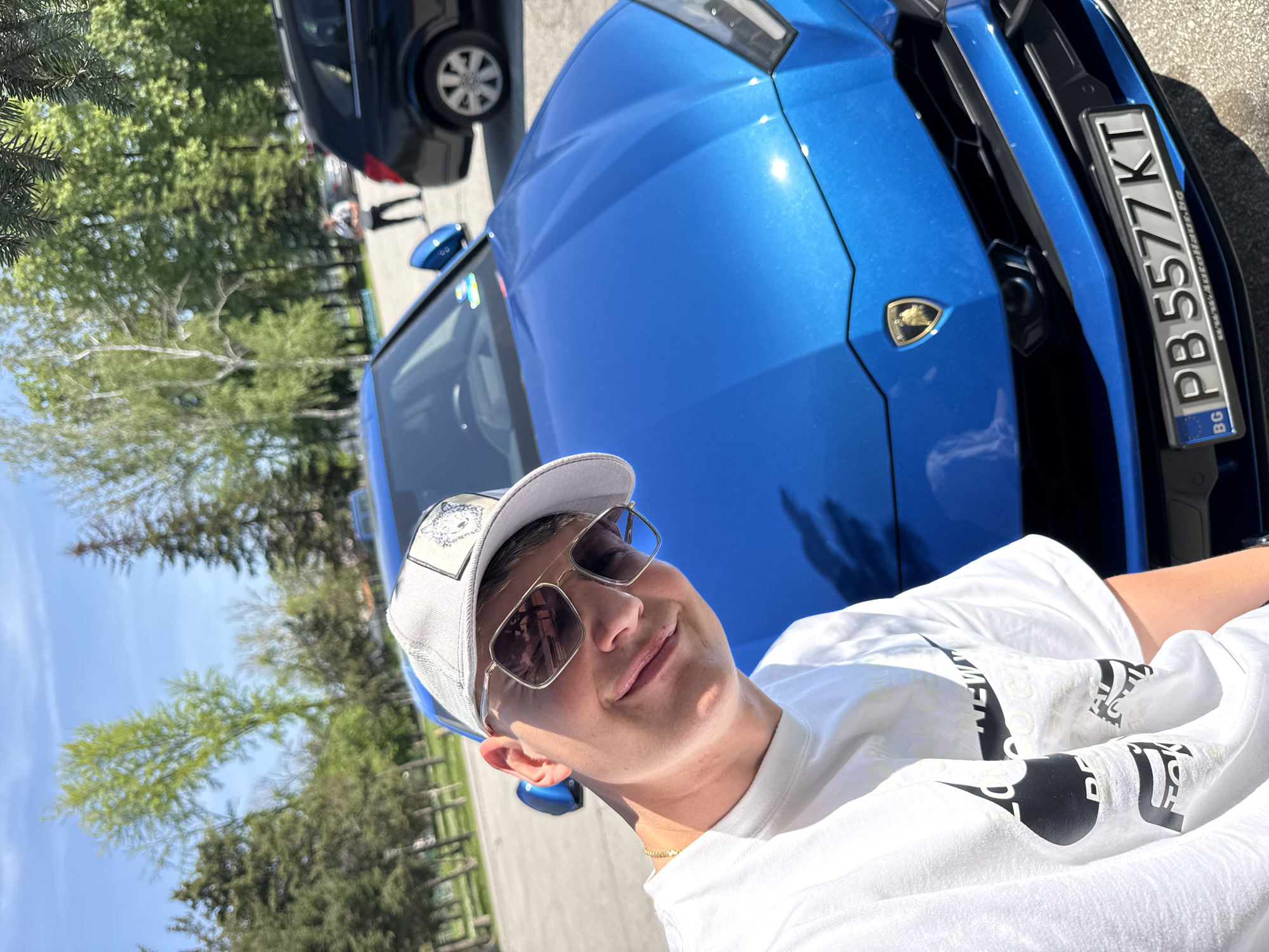 A person is seen taking a selfie outdoors, positioned in front of a bright blue Lamborghini. The image is taken at a slight angle, with the person's face dominating the foreground. They are a male-presenting individual, smiling softly at the camera, wearing a white t-shirt, a light-colored baseball cap with an animal graphic (possibly a lion or wolf head) on the front patch, and stylish square-framed sunglasses. A thin gold chain is visible around their neck.
The prominent blue car is a Lamborghini, identifiable by its badge visible on the front grille. Its metallic blue paint gleams in the sunlight, reflecting the bright sky. The front license plate of the car displays "PB 5577 KT" with "BG" (Bulgaria) indicating the country, and "www.autohaus.bg" printed below. "PB" is the regional code for Plovdiv, Bulgaria, confirming the given location context.
In the background, a dark-colored SUV or van is partially visible to the left, and further back, lush green trees and foliage are seen under a clear blue sky, suggesting a sunny daytime setting. The ground appears to be asphalt or pavement. The person's white t-shirt has some black text printed on it, partially visible, including what looks like "NEW" and other fragmented letters. The overall impression is one of leisure and enjoyment, possibly showcasing a luxury vehicle.