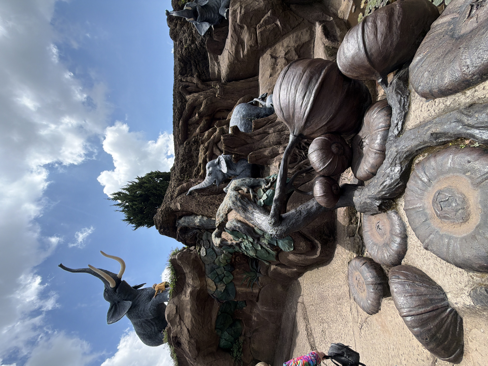 The image shows a large, dark gray elephant statue with tusks, raised trunk, and a person in a tan outfit standing on its head. This statue is positioned on a rocky outcrop with green foliage. Below, a path leads to a decorative area with large, metallic, pumpkin-like sculptures and some tree roots. In the background, there are more elephant statues on different levels of rocky terrain, surrounded by dark brown textured rocks and green plants. The sky is blue with scattered white clouds. The scene is located at the Datanla Waterfall in Da Lat, Vietnam, a tourist attraction known for its natural beauty and themed attractions.