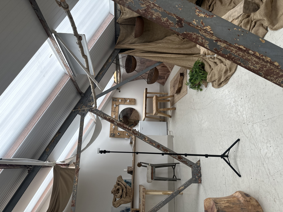 The image depicts an indoor space in London, United Kingdom, characterized by an industrial loft aesthetic combined with rustic and natural elements. The scene is illuminated by natural light filtering through translucent corrugated roof panels, which are supported by exposed grey metal trusses exhibiting significant rust and flaking paint. Tan fabric is loosely draped from parts of the ceiling structure.
In the central foreground, a black, adjustable tripod with a white, rectangular attachment on its apex stands on a light grey, possibly concrete, floor. To the left, a light wooden shelf affixed to a white wall displays a large, dark, gnarled wooden sculpture, flanked by two small wooden blocks and two framed artworks with abstract blue and white patterns. Below the shelf, a light wooden table is partially visible. A grey, backless stool with a beige cushion is positioned behind the tripod. A large, natural wood tree stump occupies the lower-left corner.
Towards the rear, a compact white mini-fridge is situated against the white wall. To its right, a rustic light wooden table is paired with a matching small wooden stool. A natural fiber rug lies on the floor in front of these, topped with a raw slice of wood. Behind this arrangement, a large rectangular wooden frame, studded with perimeter lightbulbs, encloses a circular mirror.
The right side of the space features an area draped extensively with tan, burlap-like fabric, creating a partitioned or staged section. Within this area, three dark, bowl-shaped objects are suspended by ropes from the overhead structure. On the floor in the mid-right, a cluster of green artificial foliage rests alongside additional draped burlap fabric. A bare tree branch is suspended horizontally near the ceiling structure on the right. No individuals are present in the scene, which appears set up, possibly as a studio or display area.