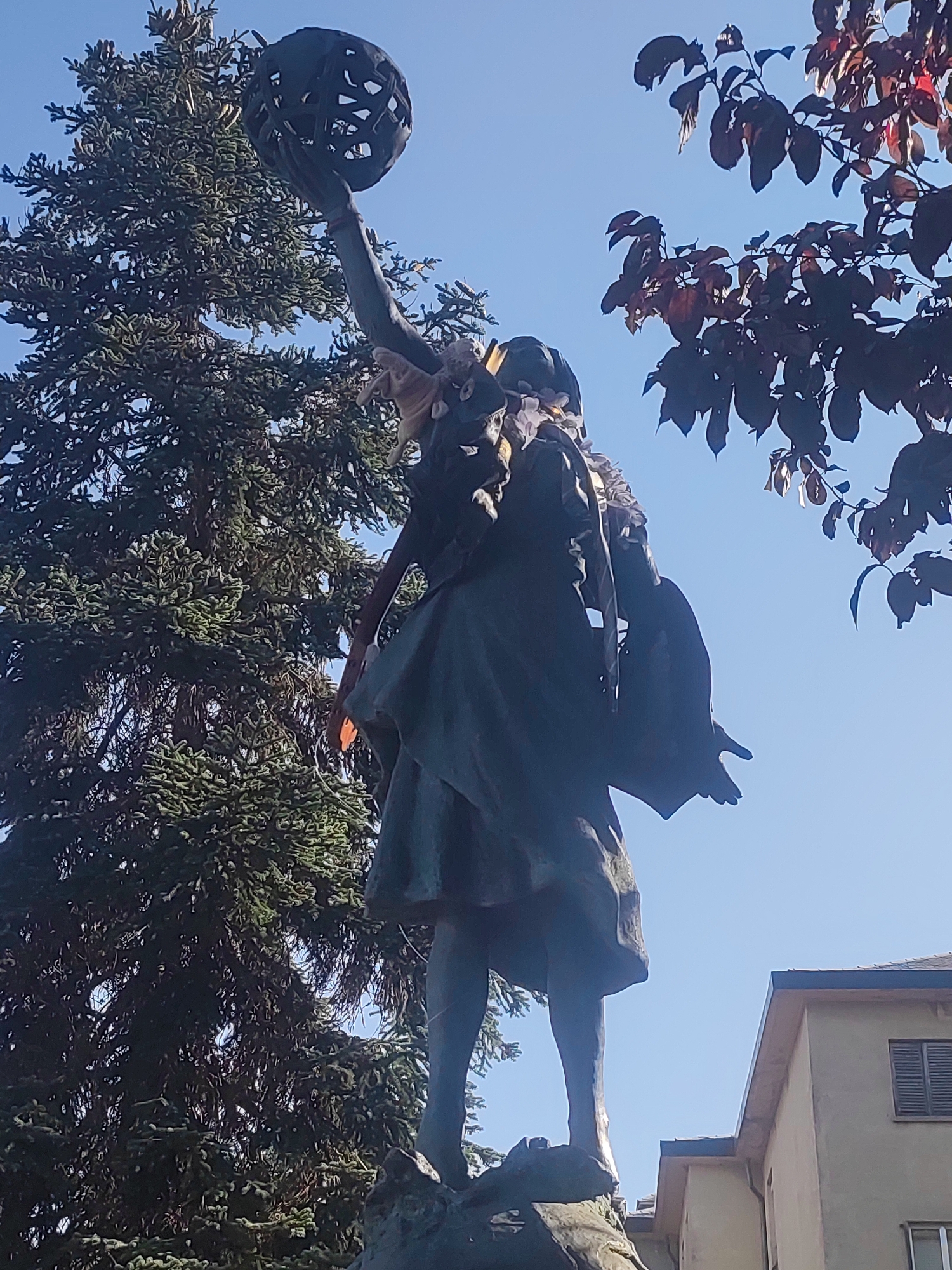 A low-angle, full-body shot captures a bronze statue of a woman holding an ornate, spherical object above her head. The statue is adorned with what appears to be a garland of flowers or leaves around her neck and shoulders, and a dark cloak draped over her left arm. Her gaze is directed upwards, and her right arm is extended, holding the sphere. The statue stands outdoors, with a large evergreen tree to the left and a tree with dark red leaves to the upper right. A building with a window and shutters is visible in the background, on the right side of the frame. The sky is clear and blue. The statue is located in the Real Sitio de San Ildefonso, Spain.