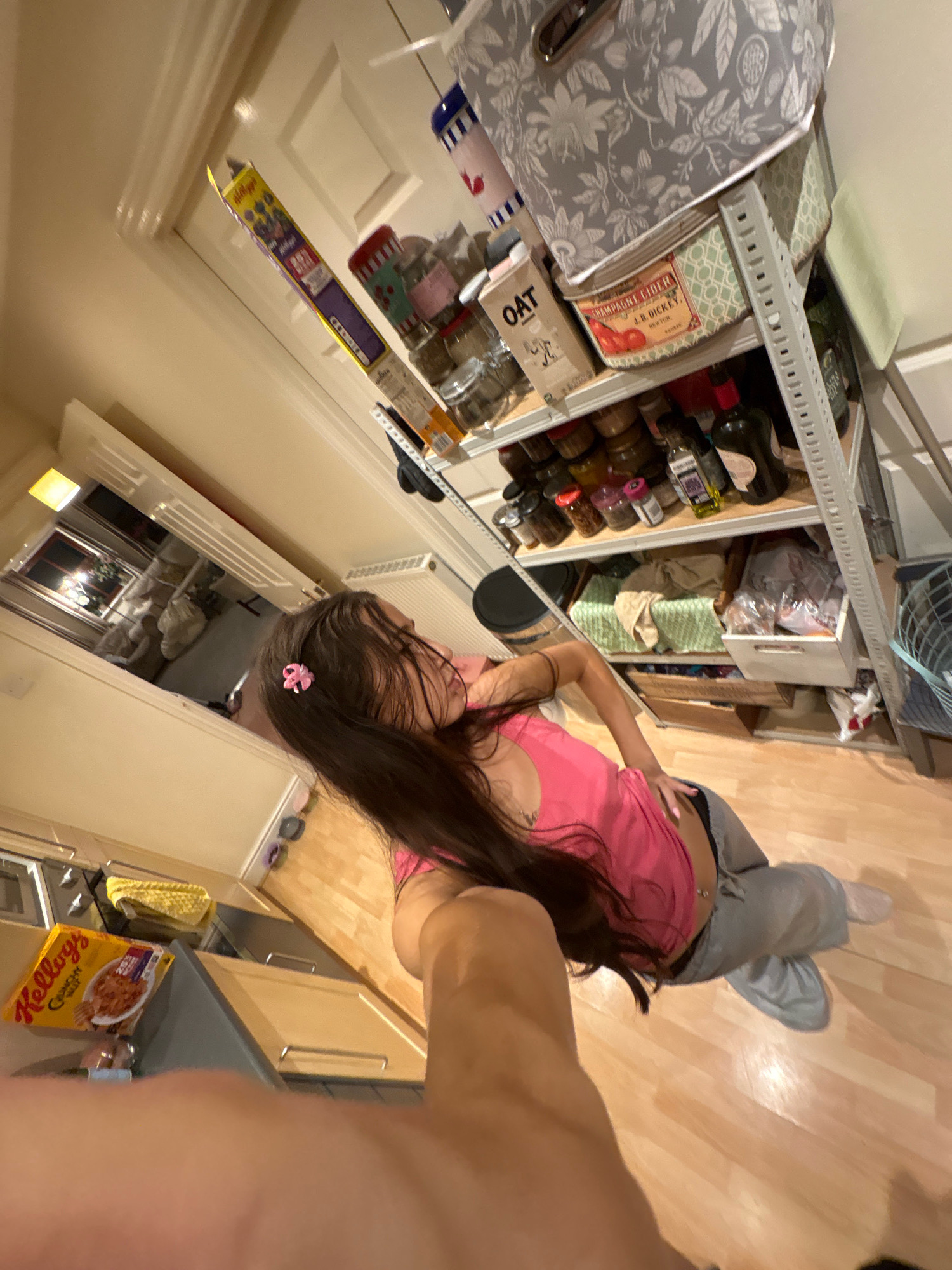 The image shows an indoor scene, likely a kitchen or pantry area. A young woman with long dark hair, wearing a pink tank top and grey sweatpants, is seated on a wooden floor. She has a small pink bow in her hair and a navel piercing. To her left, on a counter, is a yellow box of Kellogg's Crunch Nut cereal. Behind her is a multi-tiered shelving unit filled with various jars, containers, and decorative storage boxes. One prominent box on the top shelf appears to be a vintage tin with the label "CHAMPAGNE CIDER J.D. DICKEY". The environment suggests a domestic setting. The image is taken from a low, upward angle. There is no visible indication that this scene is located in Reigate and Banstead, United Kingdom.