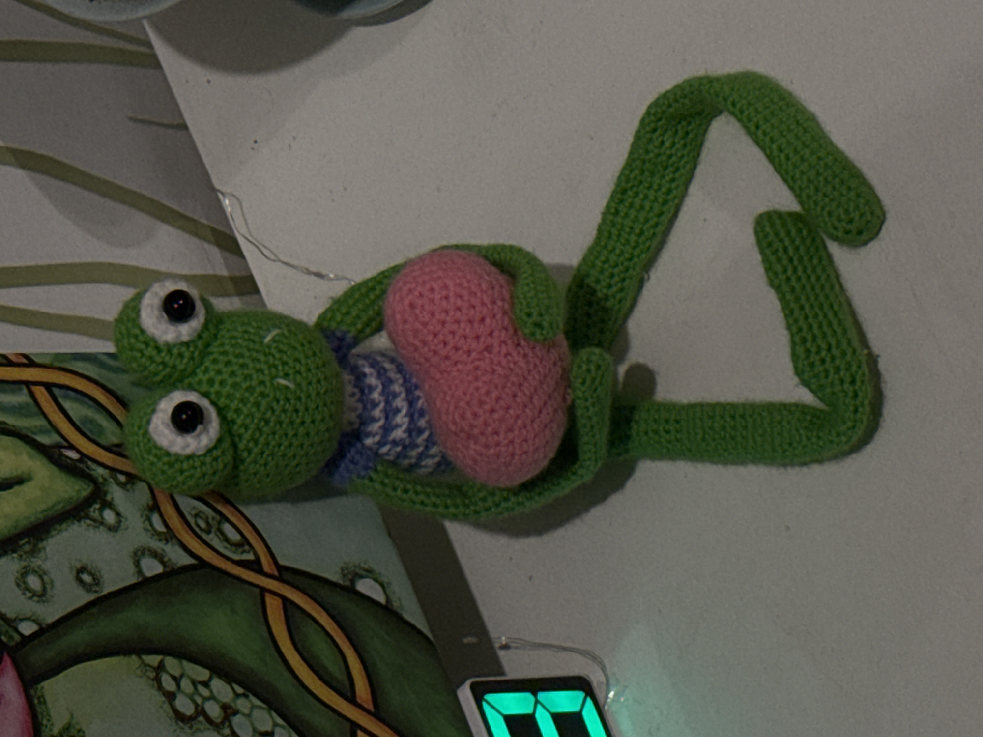 A crocheted green frog doll with white eyes, a blue and white striped shirt, and a pink heart is positioned on a white surface next to a colorful illustration and an electronic device displaying bright green numbers. The location is unknown and there is no mention of Ново-Никольское, Russia.