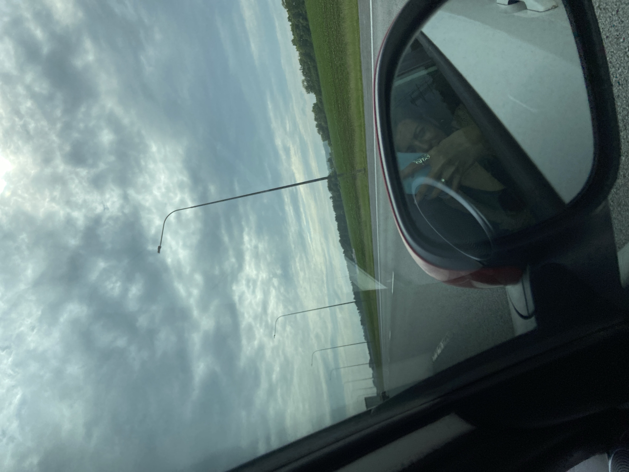 The image shows a view from inside a car, looking out the side window and into the side mirror. The sky is overcast with grey clouds. Outside the car, a highway stretches into the distance, lined with green fields and trees on either side. Several tall streetlights are visible along the highway. In the side mirror, a reflection of a person holding a phone is visible, seemingly taking the photograph. The person appears to be a woman, with dark hair, and is wearing a dark top. She is looking at the phone in her hand. The interior of the car, including part of the steering wheel and the dashboard, is also visible in the reflection. The scene appears to be a travel scene on a highway, possibly in a rural area given the presence of fields and trees. The specific location in the city of Pervomaiskoe, Russia, cannot be determined from the visual information provided in the image.