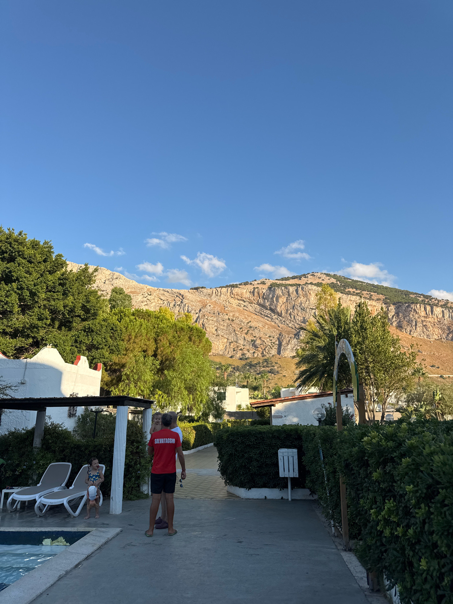 In Isola delle Femmine, Italy, an outdoor scene unfolds with a clear blue sky and scattered white clouds. In the foreground, a swimming pool is partially visible on the left. A paved walkway runs along the pool, leading towards a cluster of buildings. To the right of the walkway, lush green hedges line the path, punctuated by a white trash receptacle mounted on a pole. In the mid-ground, two lounge chairs sit empty next to the pool. A man wearing a red shirt with "SALVATAGGIO" printed on the back stands with his back to the camera, facing away from the pool. Beside him, a young child in a swimsuit is looking towards him. Further into the scene, a backdrop of trees and white buildings is visible, with a large, rugged mountain range dominating the background. The mountain's slopes are covered in sparse vegetation and appear to be rocky and striated. Palm trees and other greenery are scattered throughout the middle and background, suggesting a warm climate.