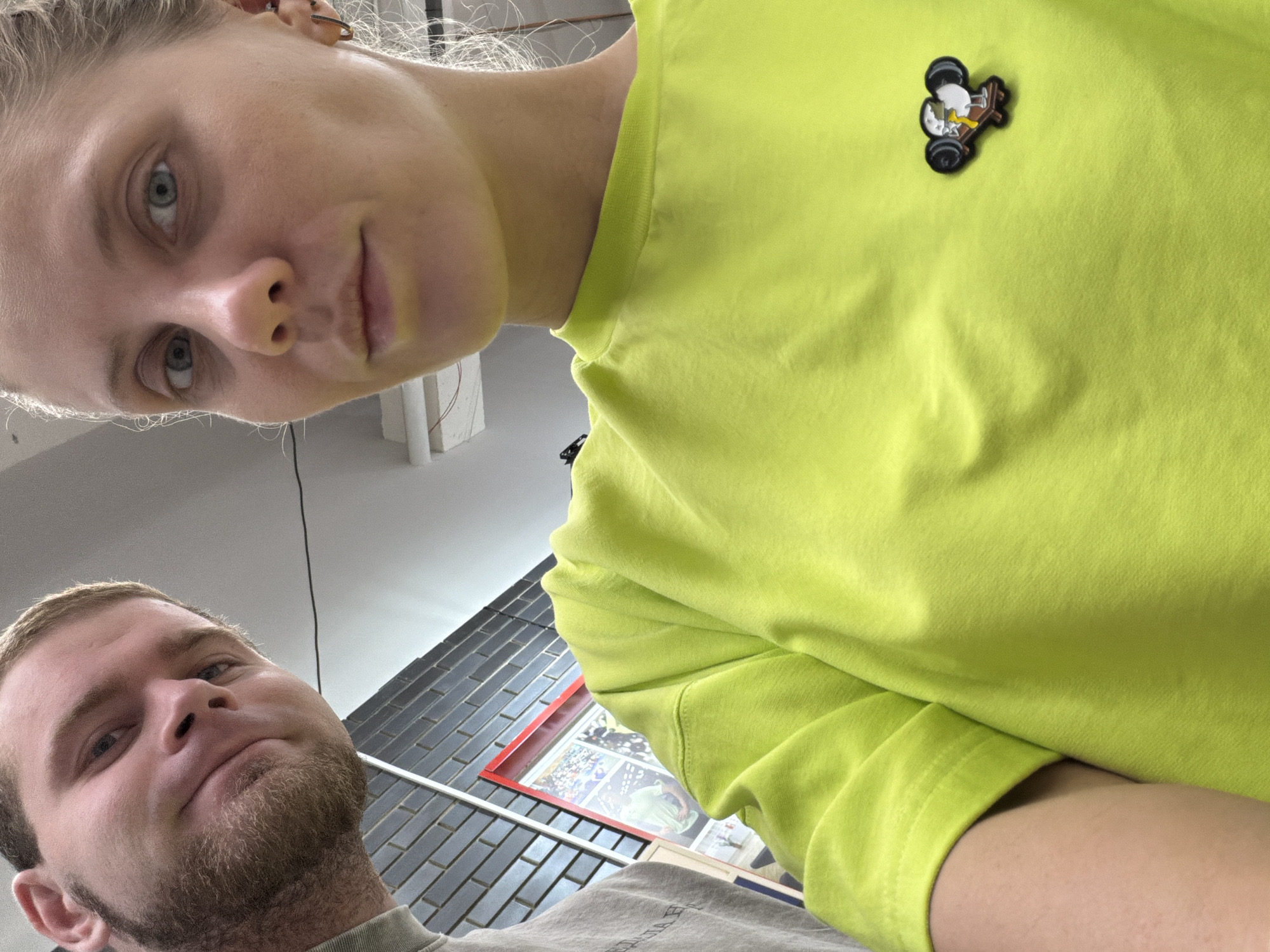 The image is a selfie of two people. A woman with blonde hair, light skin, and blue eyes is in the upper part of the frame, wearing a bright yellow-green t-shirt with a small airplane badge. A man with short brown hair, fair skin, and a light beard is in the lower part of the frame. The background shows a brick wall, a poster in a red frame, and a glimpse of what appears to be an interior setting. Given the reference point, the location is likely Chaikovsky, Russia. No actions or specific interactions are apparent other than posing for the selfie.