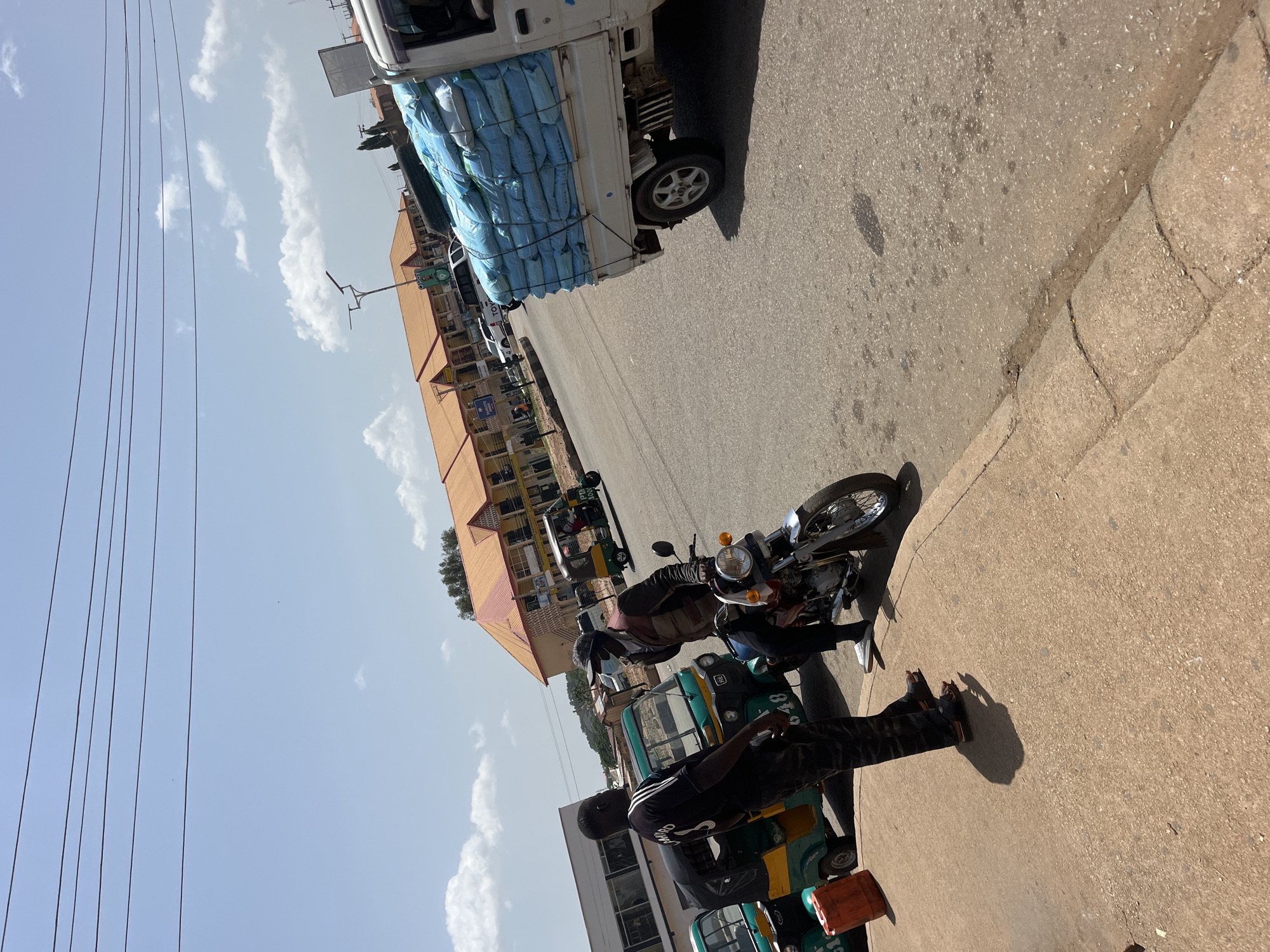 This image depicts a street scene in Gura Top, Nigeria. In the foreground, two men are interacting on a sidewalk next to a parked motorcycle. One man, wearing a black shirt with white stripes and black pants, stands facing away from the camera, looking towards the other man. The second man, wearing a dark vest over a lighter colored shirt, a cap, and a face mask, is seated on a motorcycle. The motorcycle is positioned on the road, adjacent to the sidewalk.
Behind the men, several auto-rickshaws, also known as Keke Napeps, are parked along the side of the road. One auto-rickshaw is a bright green and yellow, with the number "048" visible on its side. Another is partially visible behind it. Further down the street, a white truck is driving by, heavily loaded with large, blue sacks secured with ropes.
The background features buildings with distinctive orange-tiled roofs, suggesting a commercial or residential area. Power lines crisscross the sky, and a street lamp is visible. The sky is a pale blue with scattered white clouds, and the sun appears to be casting shadows. The overall impression is of a busy street in a Nigerian city.